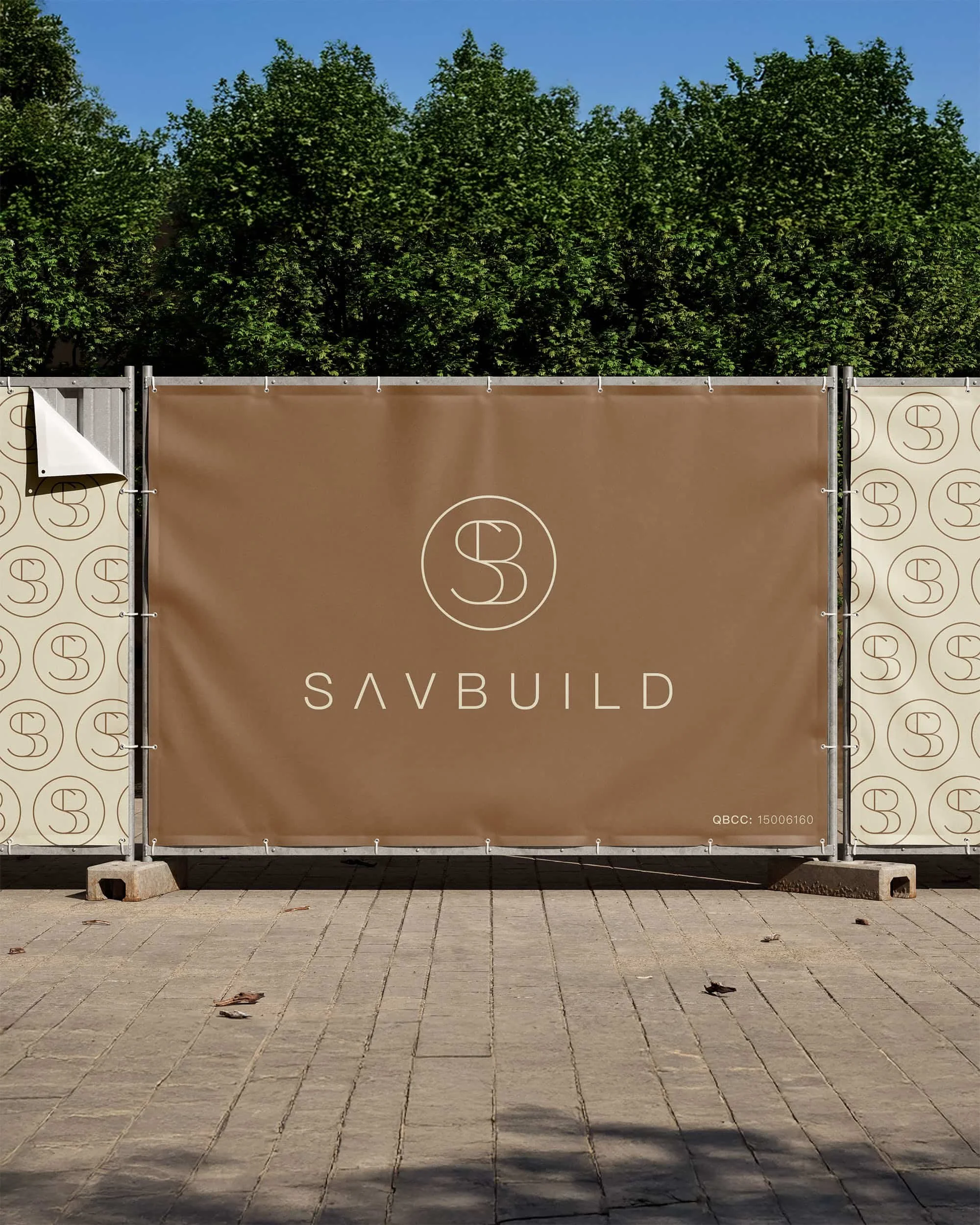 Construction site fence panel displaying a logo and the word 'SAVBUILD' under a blue sky with green trees in the background.