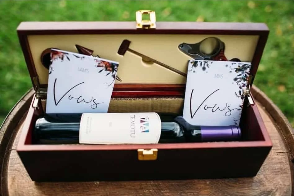 A wooden box contains a rolled wine bottle, two note cards labeled 'Vows' for June and March, a black and green wine opener, a wine stopper, and a purple-capped wine bottle opener, set outdoors on a wooden surface.