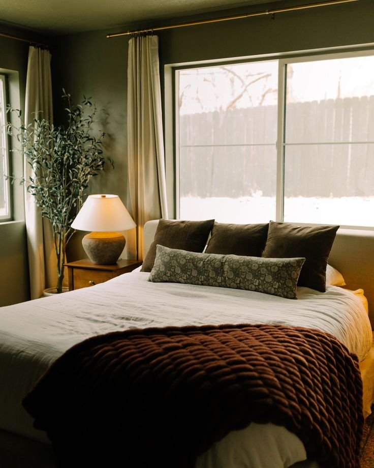 A cozy bedroom with a large bed covered in white bedding and a textured dark brown blanket. The bed has decorative pillows. To the left, there’s a wooden nightstand with a beige table lamp. A tall green potted plant stands in the corner.