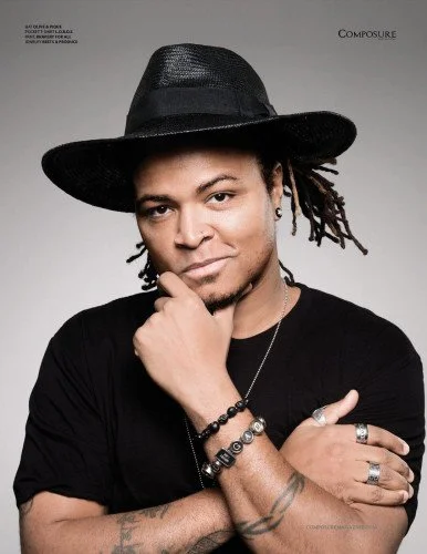 A man wearing a black wide-brimmed hat, black T-shirt, and various bracelets and rings, posing with his hand on his chin and looking directly at the camera.