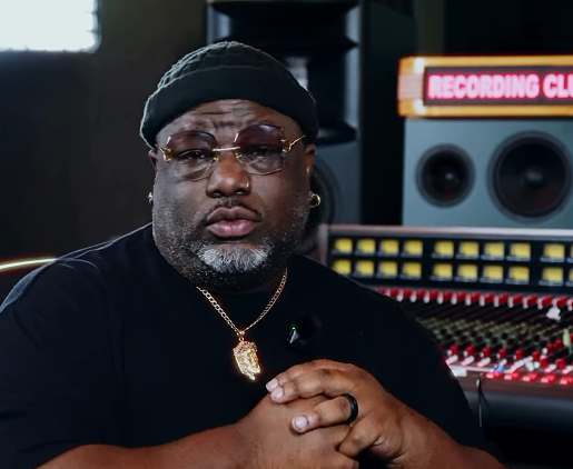 A man wearing a black beanie, glasses, and a gold chain, sitting in a recording studio with audio equipment in the background.