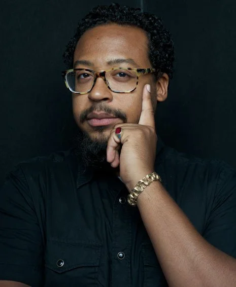 A man with curly hair, glasses, and a beard, wearing a black shirt and a gold bracelet, touching his temple with his index finger against a dark background.