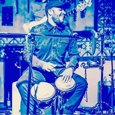 A man playing bongos and singing into a microphone on stage, wearing a dark shirt and cap, with a musical setup in the background.