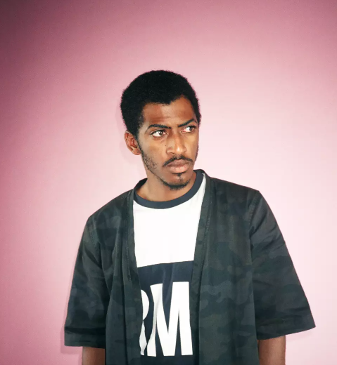 Man with short curly hair and a beard looking to the side, wearing a black jacket over a white t-shirt with black lettering, against a pink background.