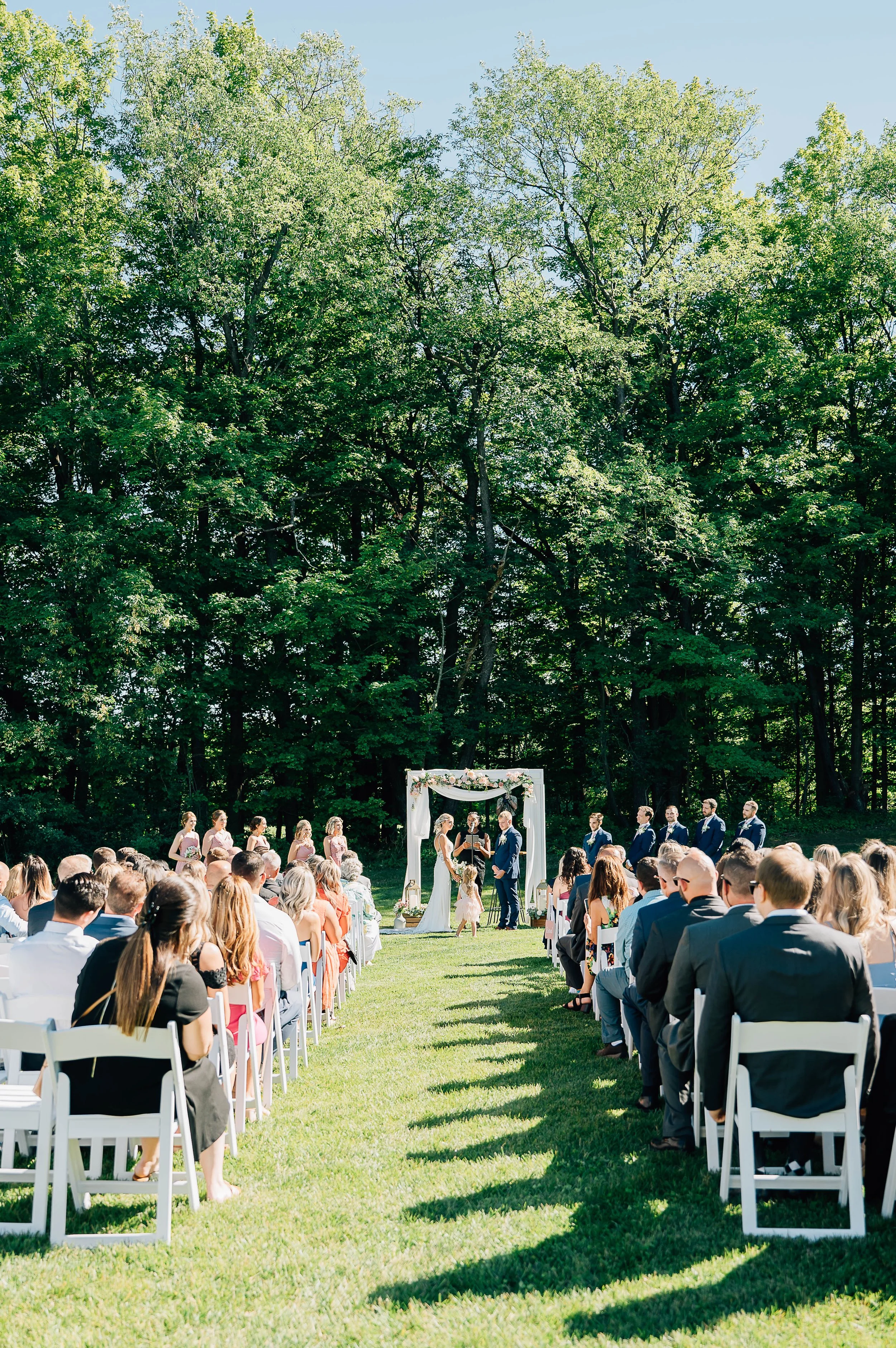Outdoor wedding ceremony at secluded farm wedding venue with wooded background white arbor and white ceremony chairs in Buffalo NY