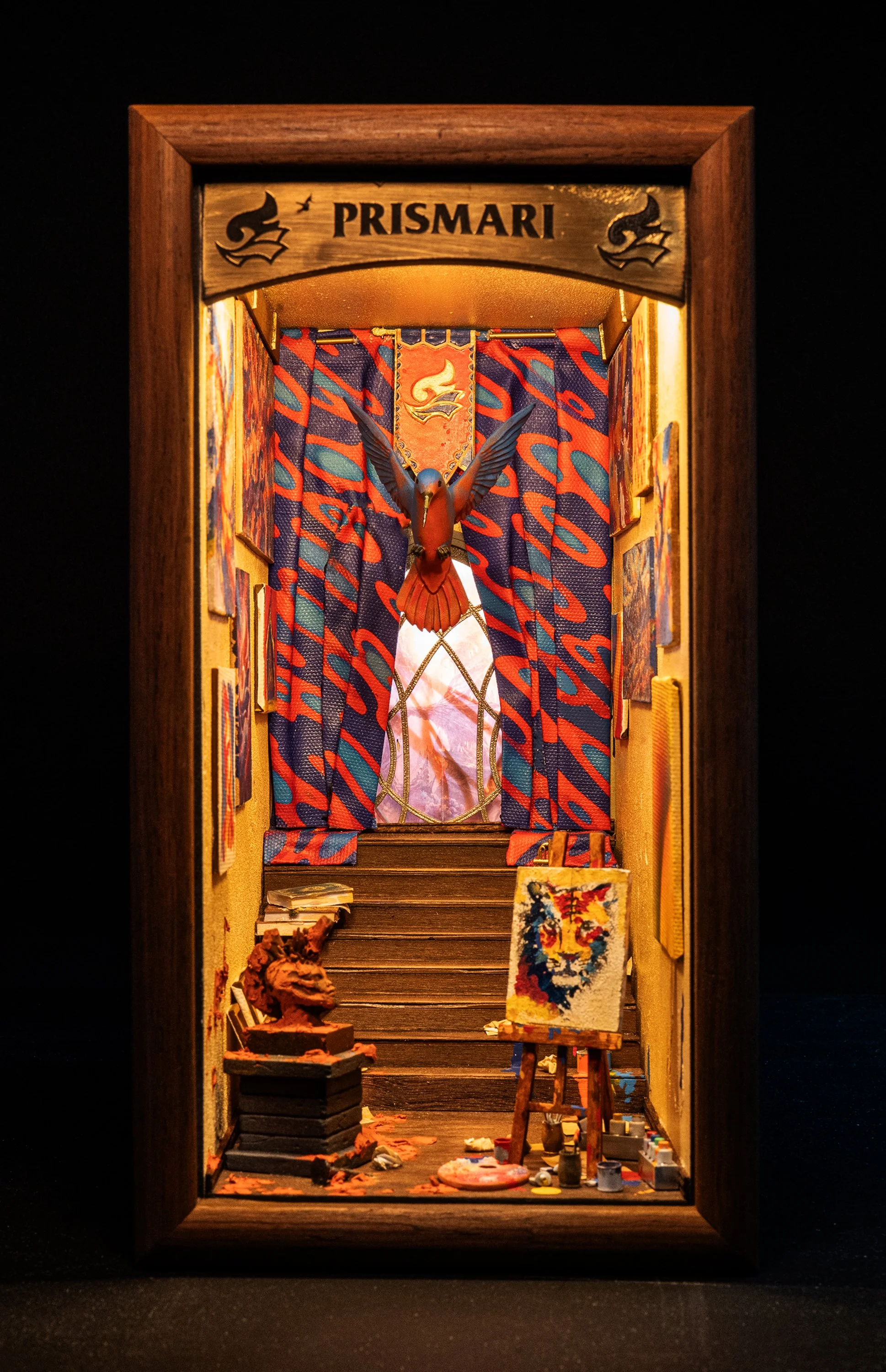 Miniature art studio scene with wooden frame, colorful fabric curtains, a bird sculpture in the background, an easel with a painted canvas, art supplies and sculptures on the floor, and the word 'PRISMARI' on a wooden sign at the top.