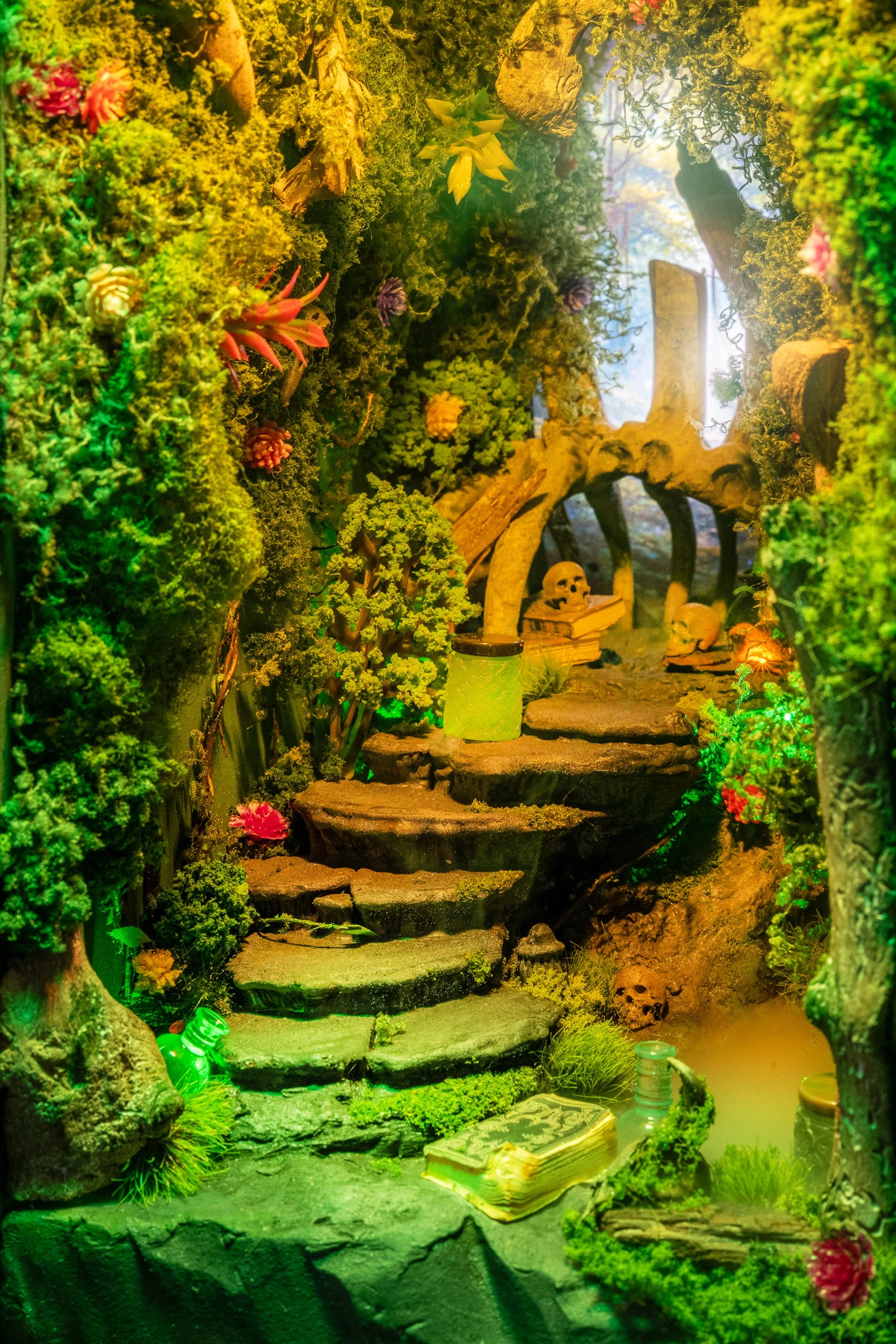 A miniature fantasy scene of a lush, moss-covered forest with stone steps leading to a small water feature, decorated with colorful plants, skull ornaments, and a glowing green bottle in the foreground.