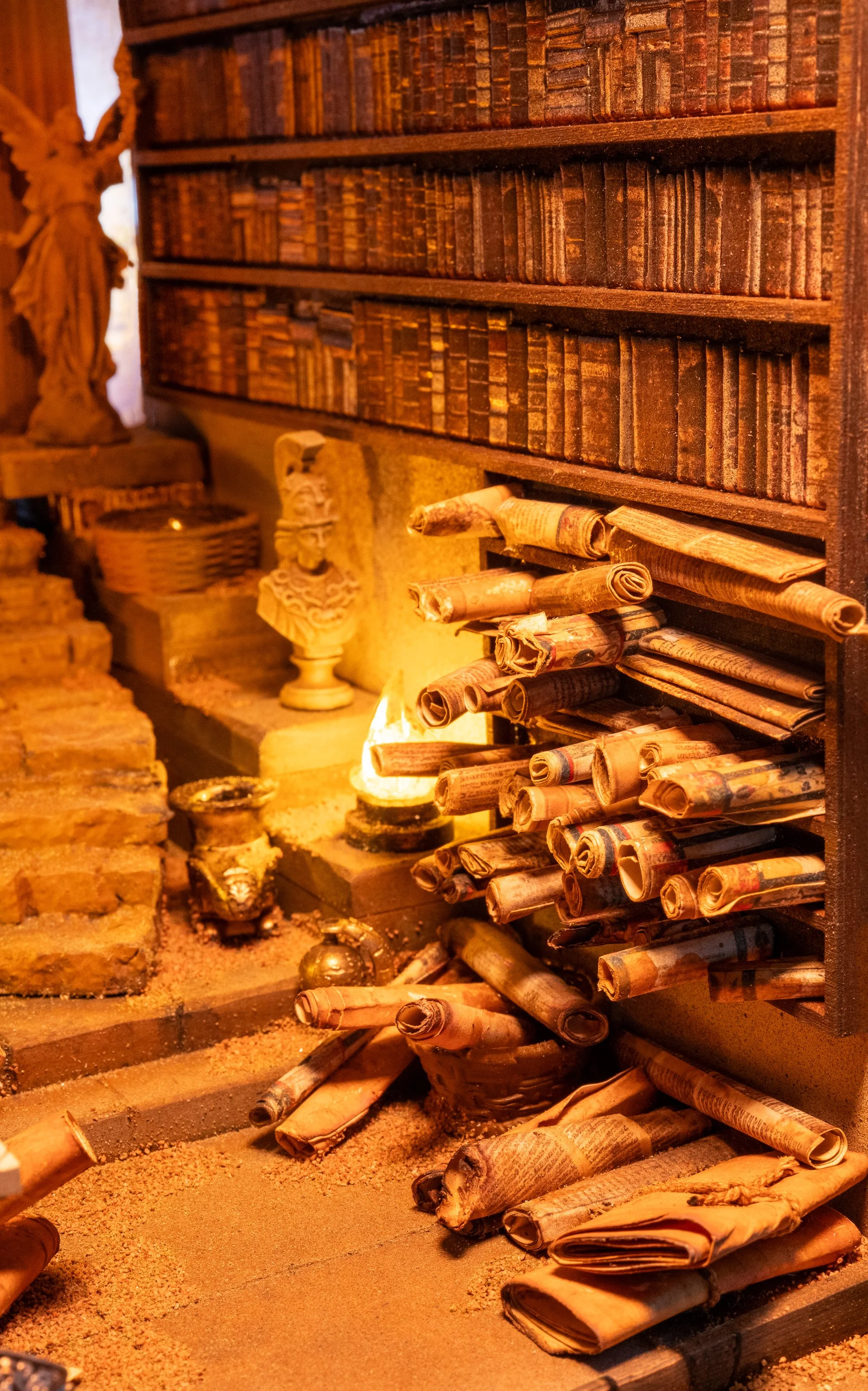 A cozy library scene with a large wooden bookshelf filled with old books, scrolls, and rolled-up newspapers. There are decorative artifacts, a lit oil lamp, and a statue of an angel, creating a warm, vintage ambiance.