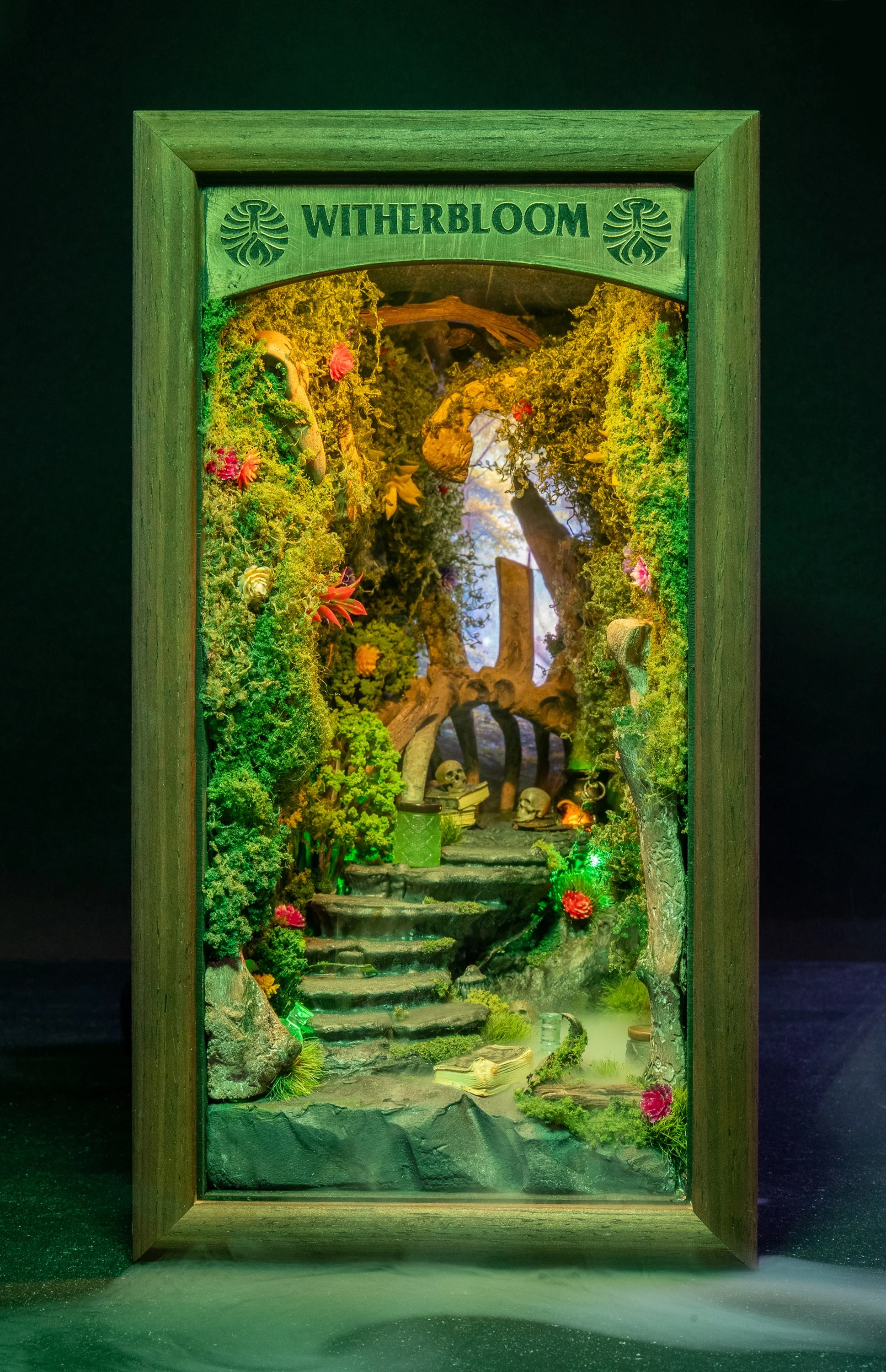 A miniature waterfall scene within a wooden frame labeled 'WITHERBLOOM', featuring lush green moss, colorful flowers, small skulls, a staircase, and miniature books, illuminated with green and yellow lighting to create a mystical forest atmosphere.