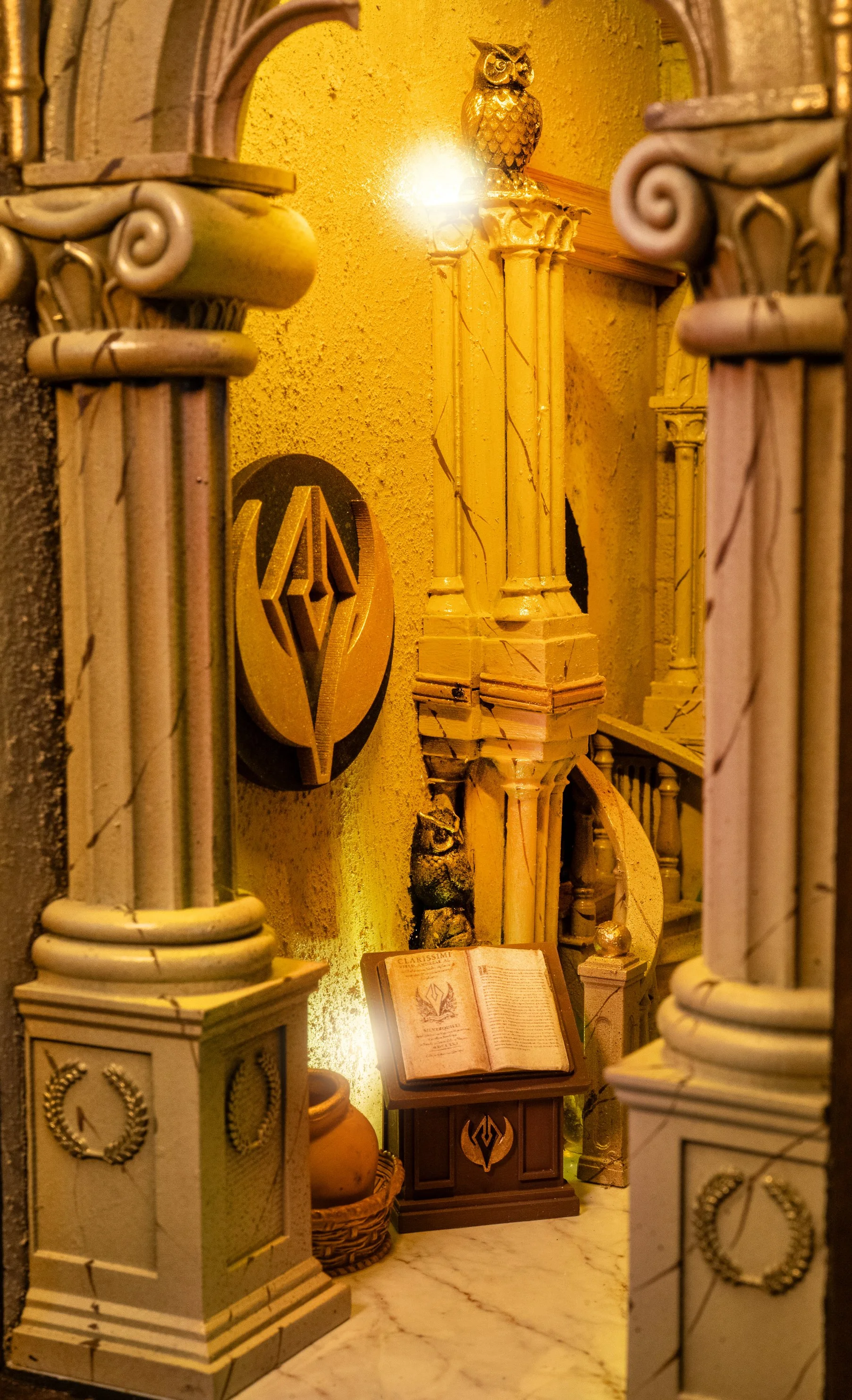 A decorative scene featuring ornate white columns with gold accents, an open book on a small table, a ceramic pot, a wooden stand with a book, a golden owl figure on a pedestal, and a gold and black emblem on the wall, all illuminated by warm yellow 