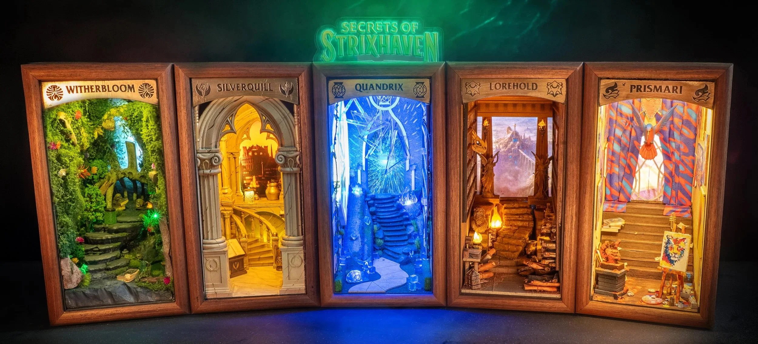 A wooden display with five miniature dioramas labeled Witherbloom, Silverquill, Quandrix, Lorehold, and Prismari, each depicting fantasy-themed scenes with vibrant lighting and detailed scenery.