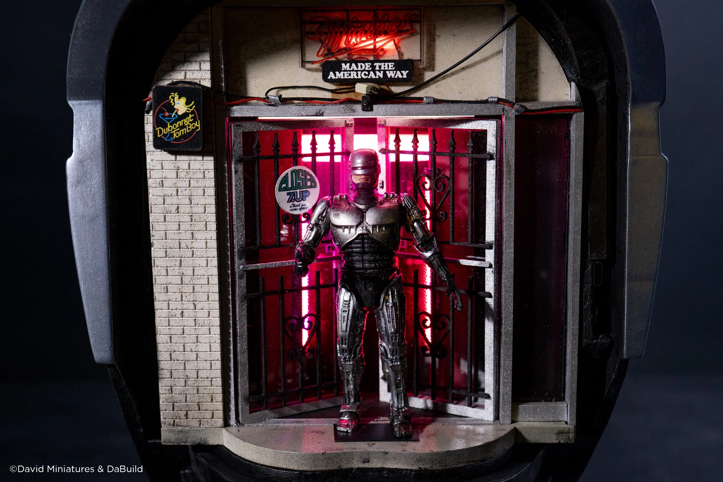 A detailed miniature diorama of a robot standing in front of a small building with neon signs. The building has a brick wall exterior and signs that read 'Made the American Way,' 'Closed,' and 'Dubonet TomBoy.' The robot has a metallic, robotic appea