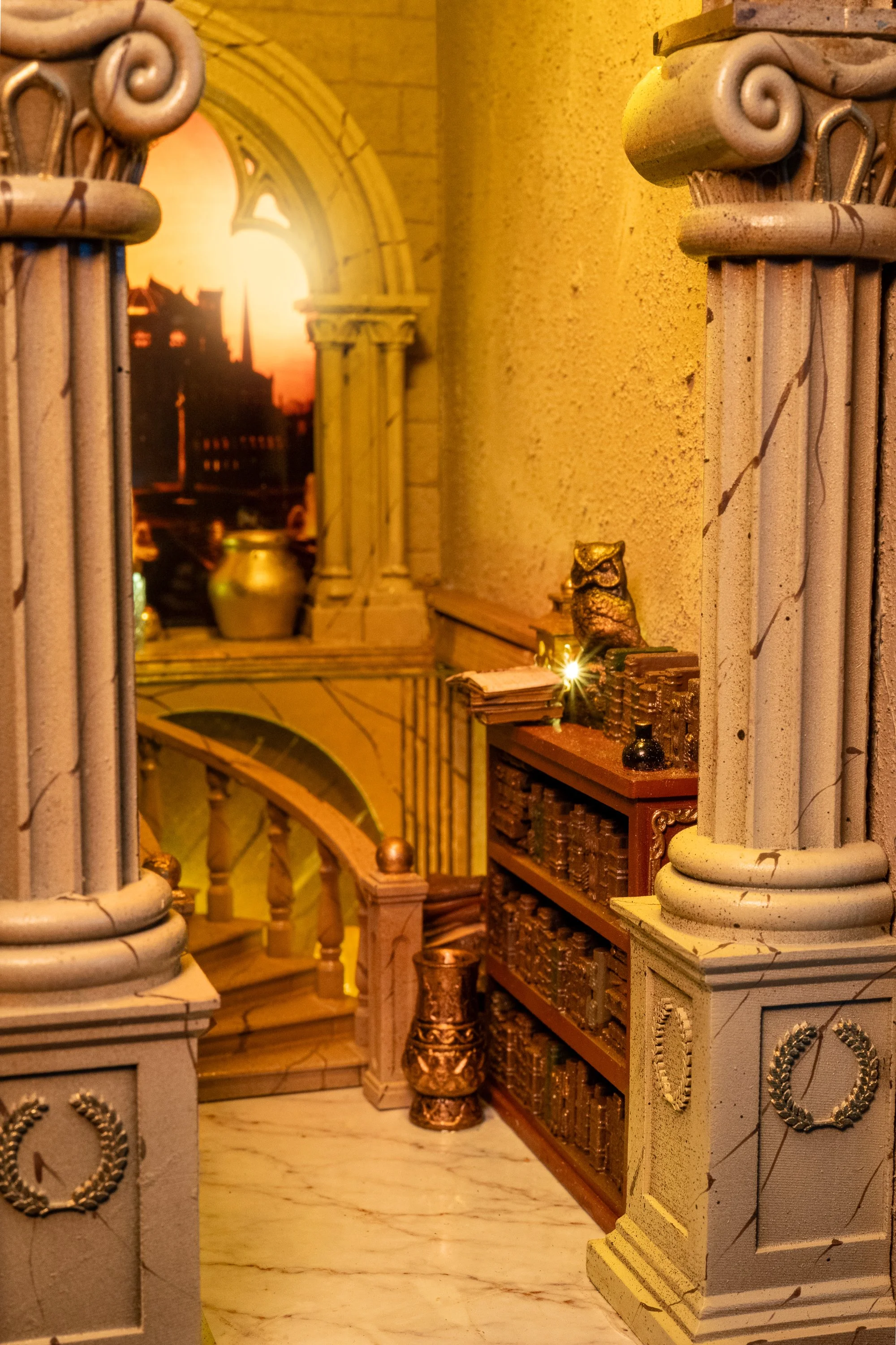 Interior of a building with ornate columns, a staircase, decorative gold and bronze objects, and a large window with a sunset view of a city skyline.