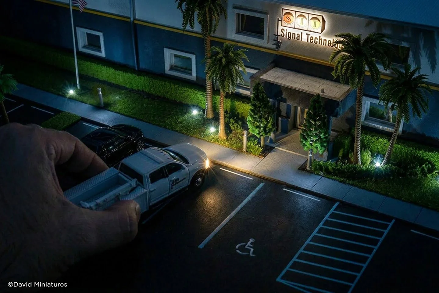 Miniature model of a parking lot with a handicap space, a chair, parked cars, a sidewalk, palm trees, and a building with a sign that reads 'Signal Tech'.