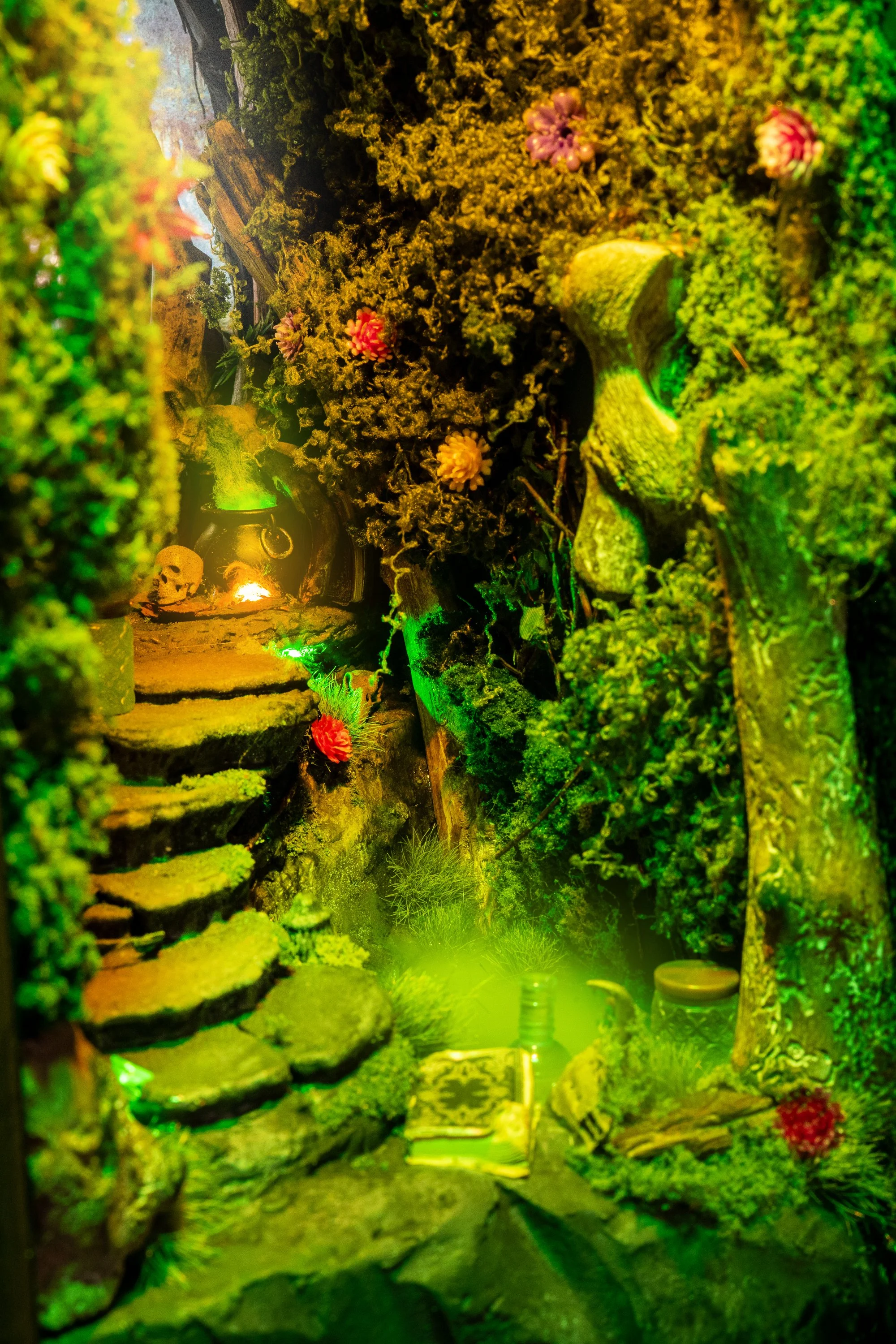 A miniature fantasy scene with moss-covered walls, colorful flowers, stone stairs, and a small glowing skull near a river, illuminated by green and yellow lights.