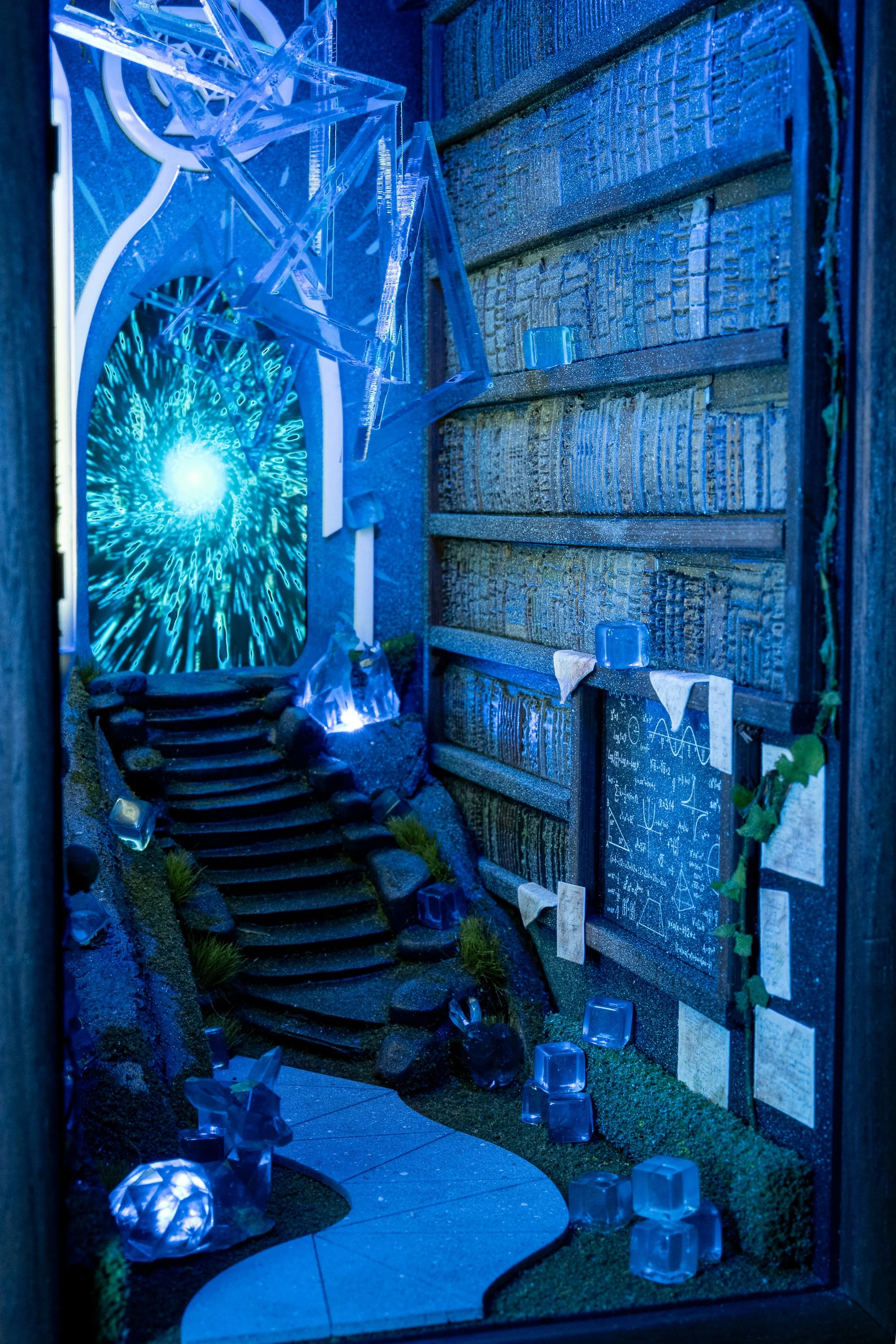 A miniature artistic outdoor scene illuminated with blue lighting, featuring a staircase made of rocks, a winding pathway, a blackboard with mathematical formulas, a small greenhouse, a spherical sculpture, and several glass blocks surrounding the sc