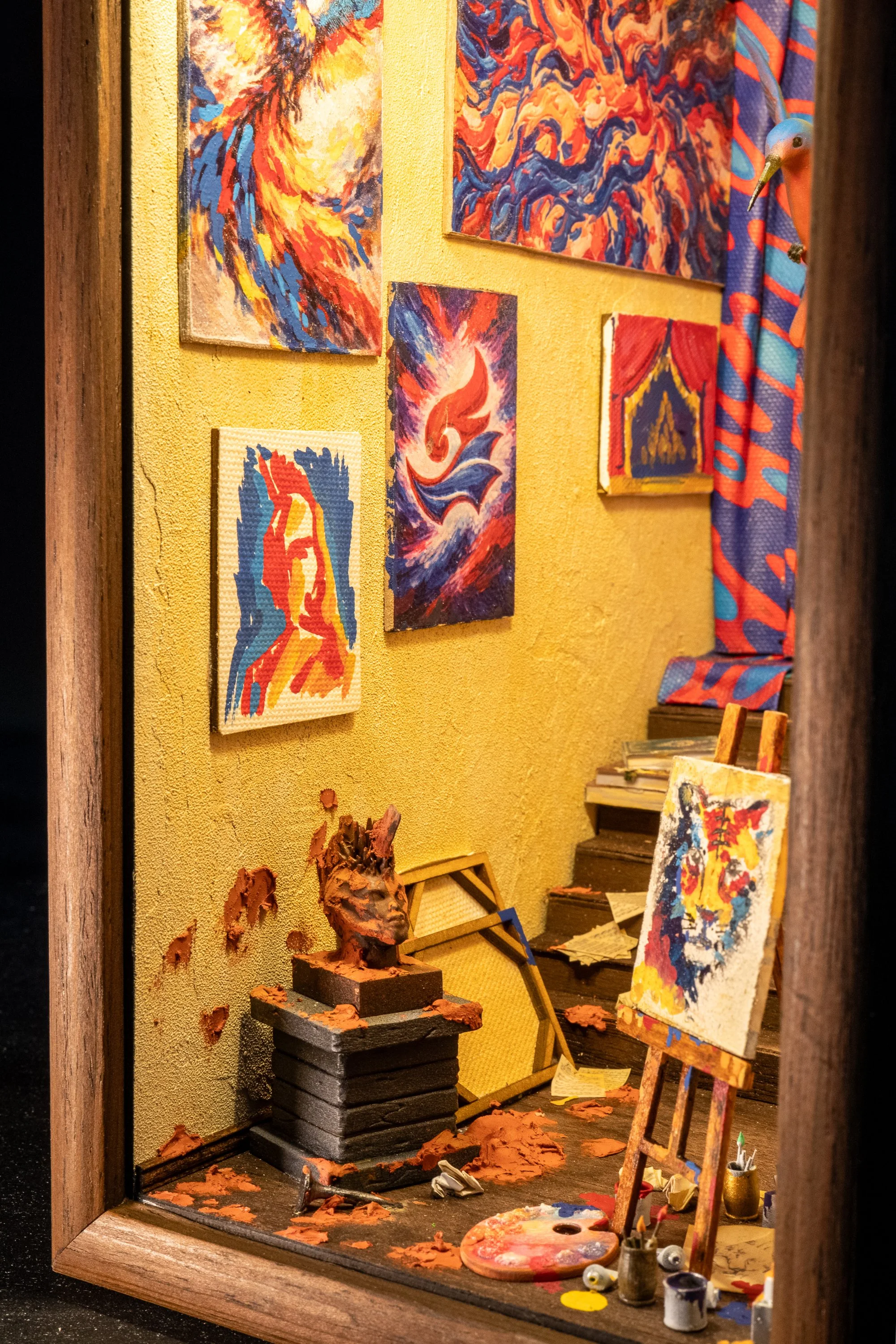 A miniature art studio diorama featuring wall-mounted paintings, a wooden easel with a colorful abstract painting, art supplies including brushes, cans of paint, a palette, and a clay sculpture on a black pedestal. The background has yellow textured 