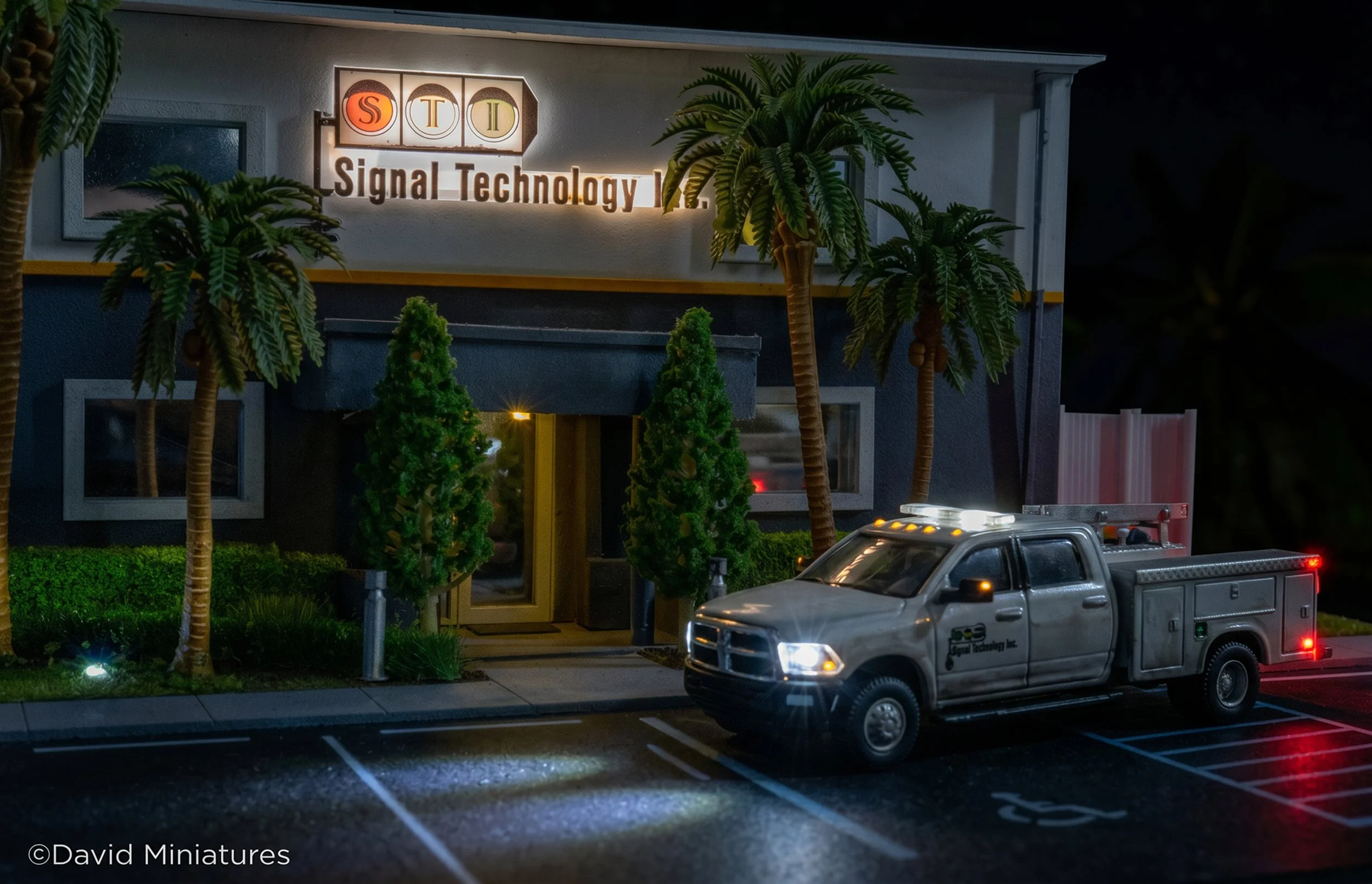 Miniature model of a building with a sign that reads "Signal Technology" and a logo with the letters 'S T I'. The scene includes miniature trees, a model vehicle, and parking lot details, set against a night background.
