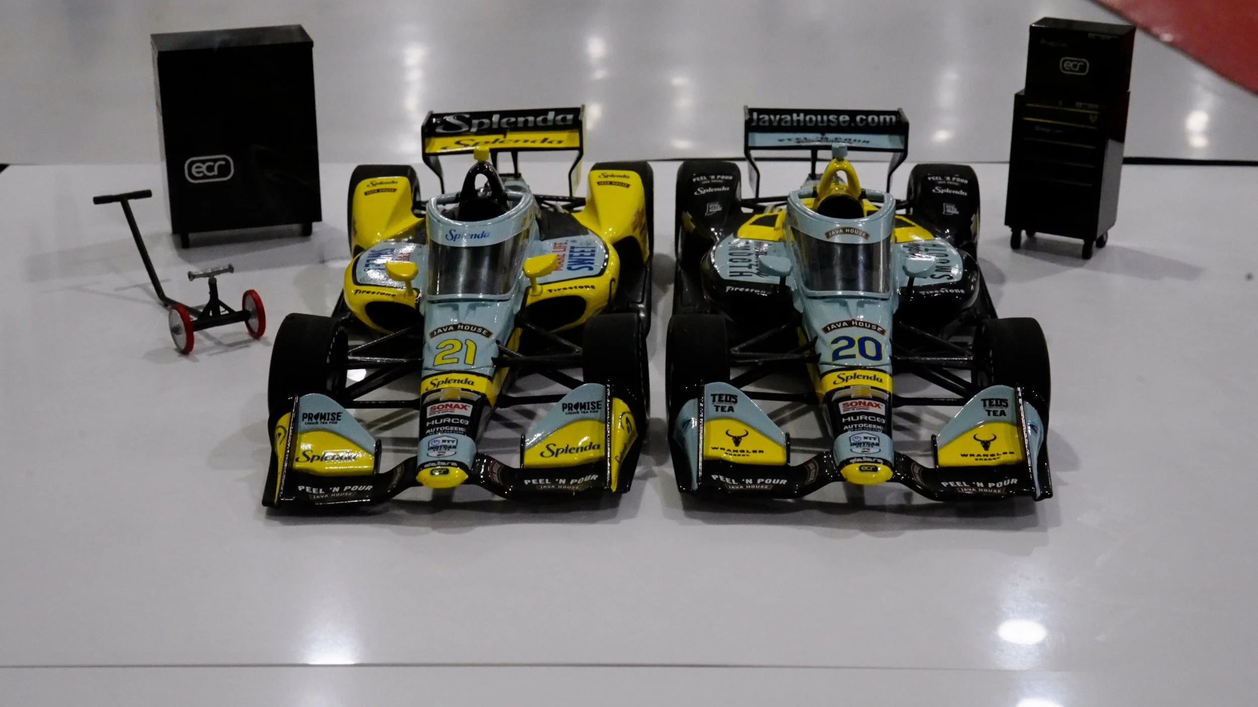 Two miniature racing cars with yellow, blue, and black livery, parked side by side on a white surface, with a small red and black pushcart and black storage cabinets in the background.