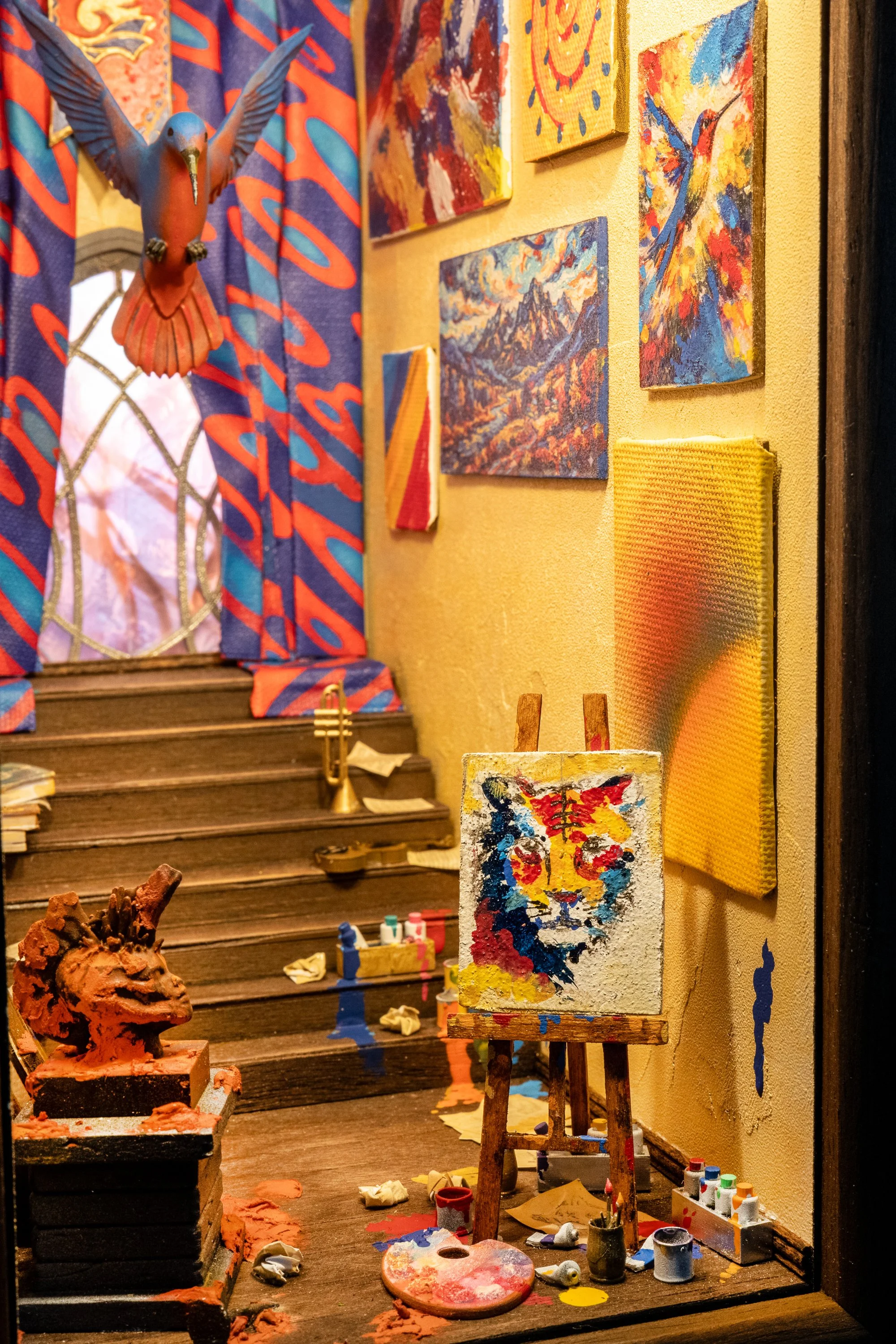 An art studio with colorful paintings displayed on a beige wall, an easel with an abstract painting, various art supplies scattered on a wooden surface, and a decorative bird sculpture hanging near a window with blue and red curtains.