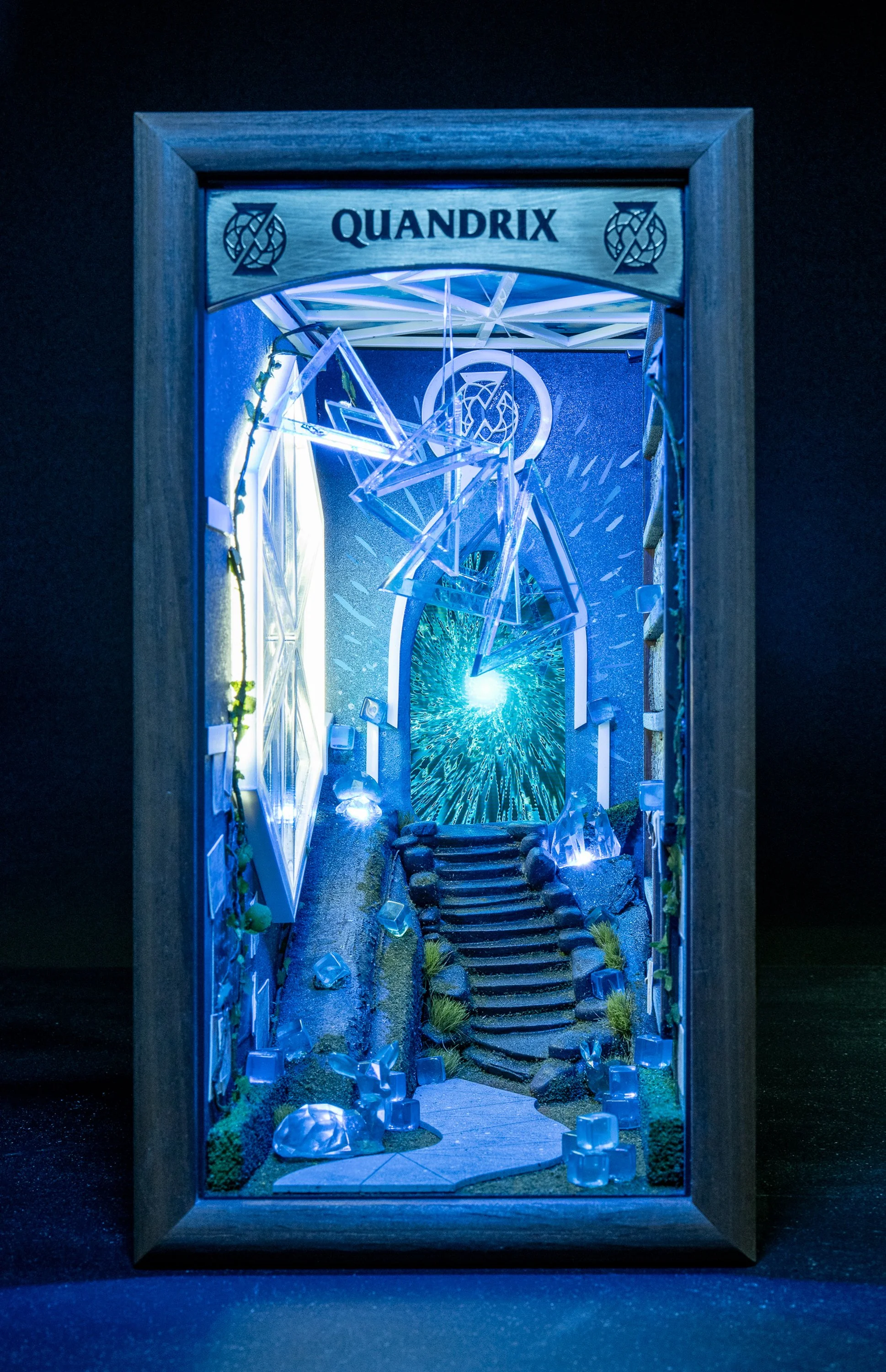 Art installation titled 'QUANDRIX' featuring illuminated geometric shapes, mirrors, and staircase within a wooden frame, creating a futuristic scene with blue lighting.