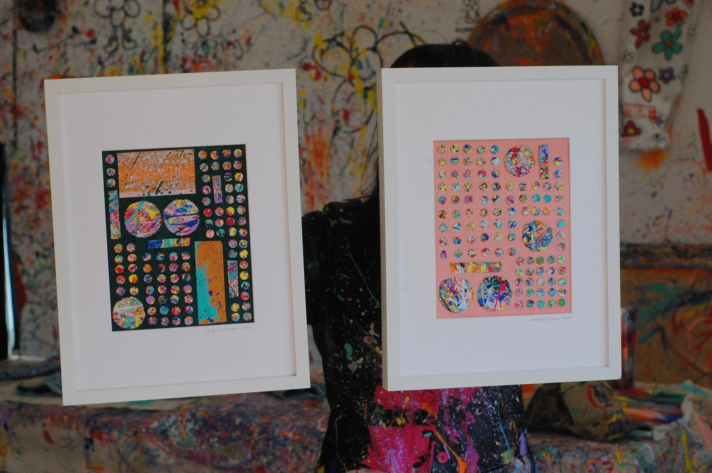 Artist holding two framed SUPERBLOOM artworks—Repurpose No. 2 on deep green linen and a second pink upcycled piece—inside a colorful paint-splattered studio.