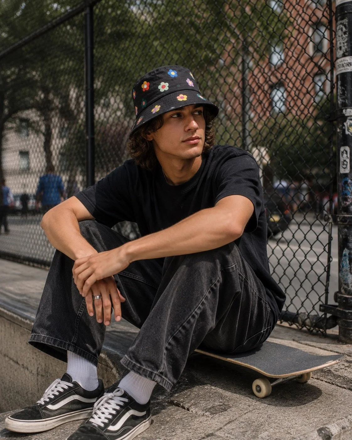 A skater sits on a skateboard in an urban park wearing a black bucket hat decorated with colorful five-petal flowers and relaxed streetwear.