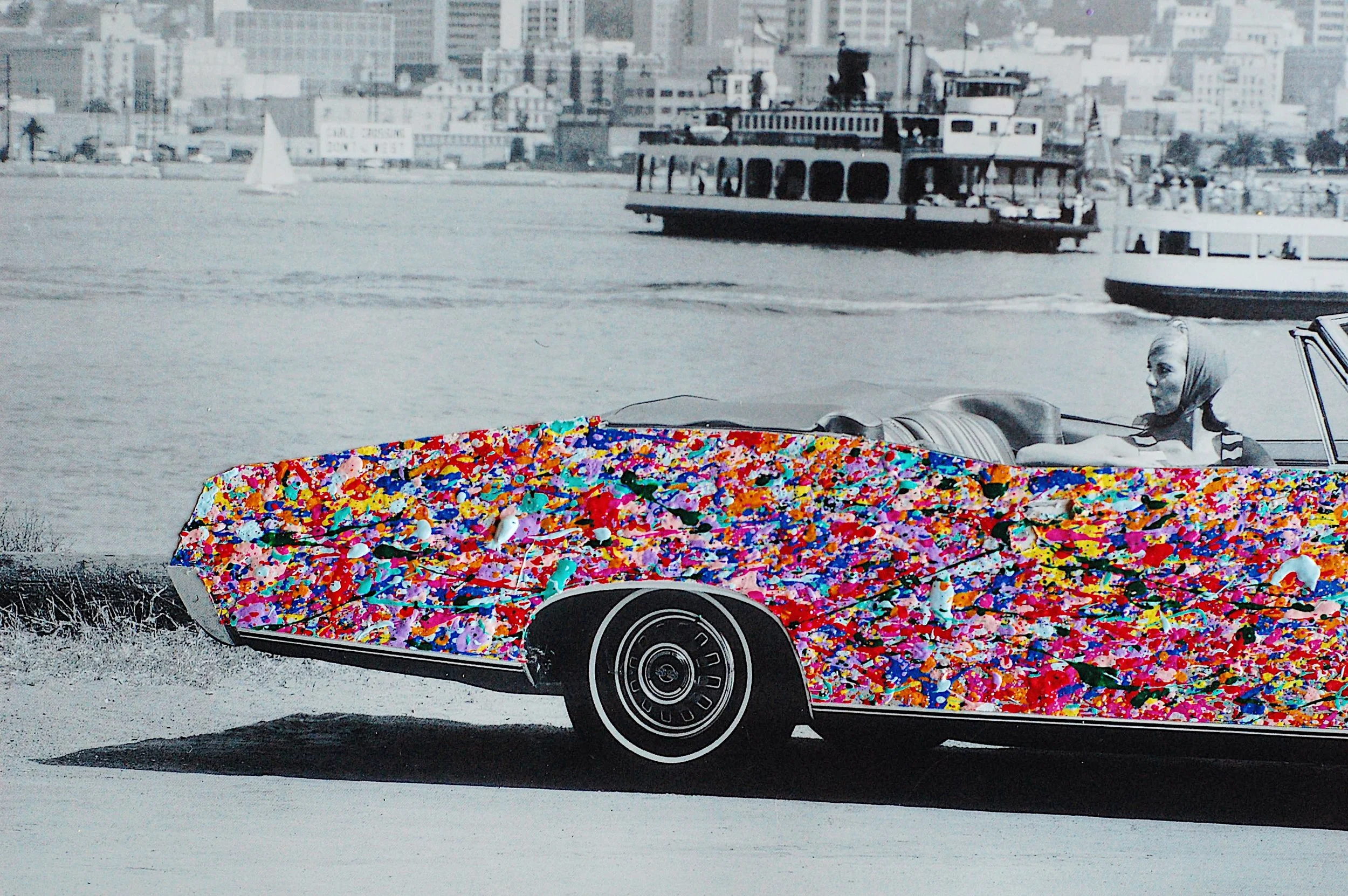 Close-up of a vintage convertible driver with the car’s body covered in layered, colorful paint splatter against a grayscale harbor background.