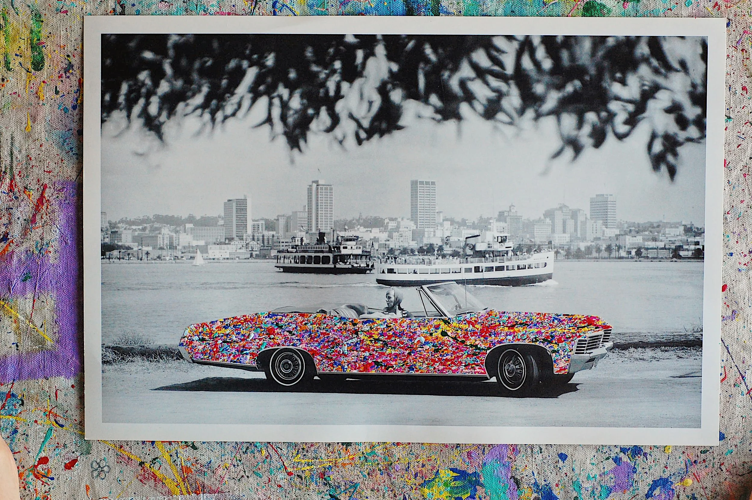 Vintage black-and-white photograph of a 1967 Chevrolet Impala convertible overlaid with vibrant multicolor paint splatter, viewed from above on a paint-covered studio surface.