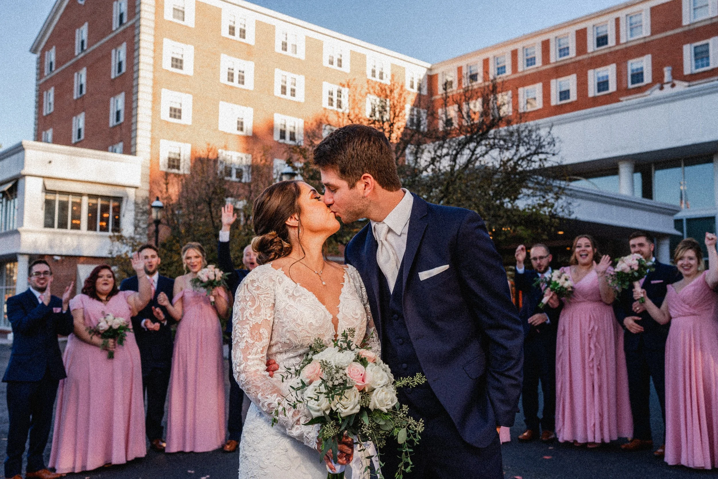 A newlywed couple sharing a kiss outside a city building during their wedding celebration, with friends and family cheering in the background.