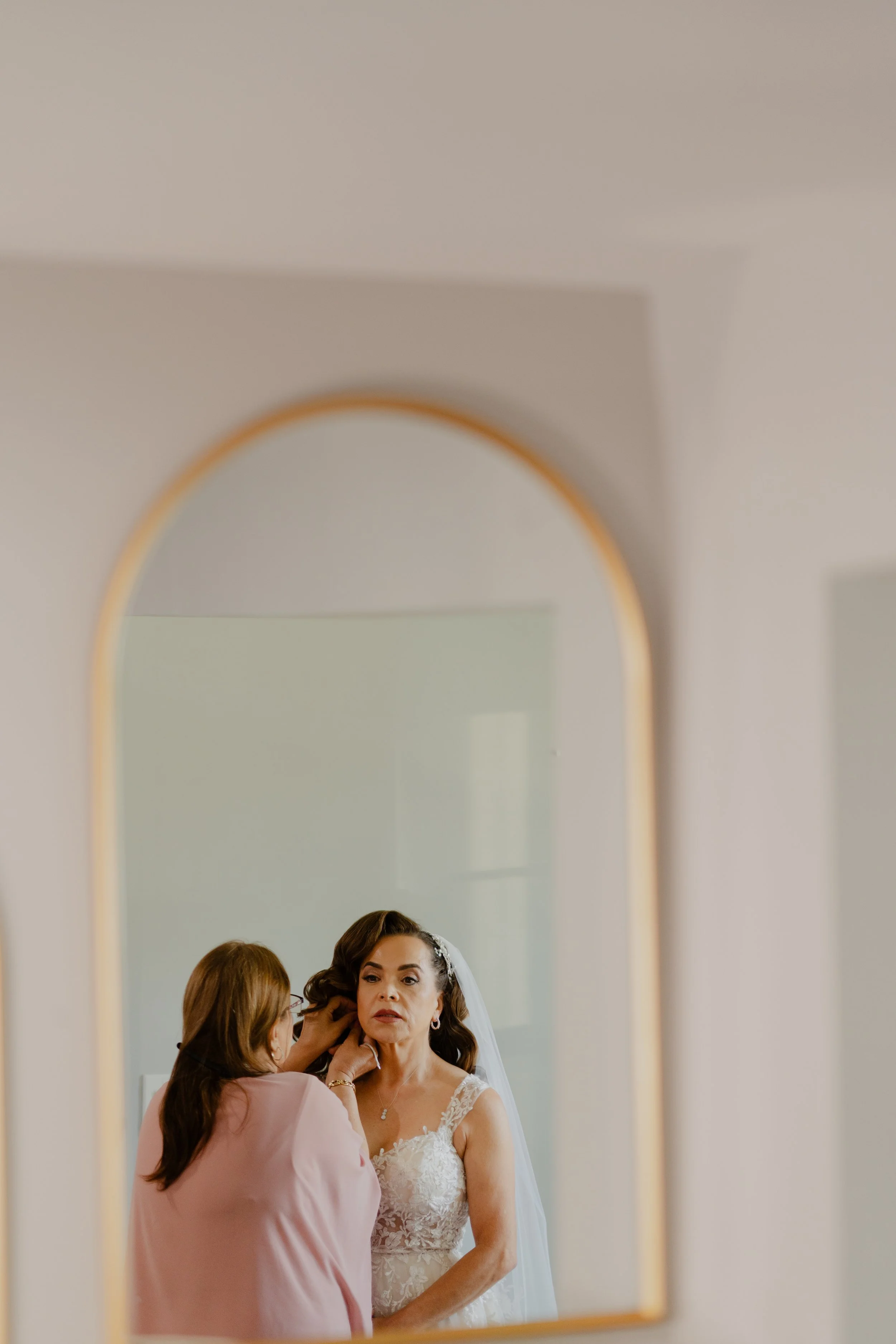 A woman in a wedding dress is having her makeup or hair done in front of a mirror, with a person adjusting her earrings.