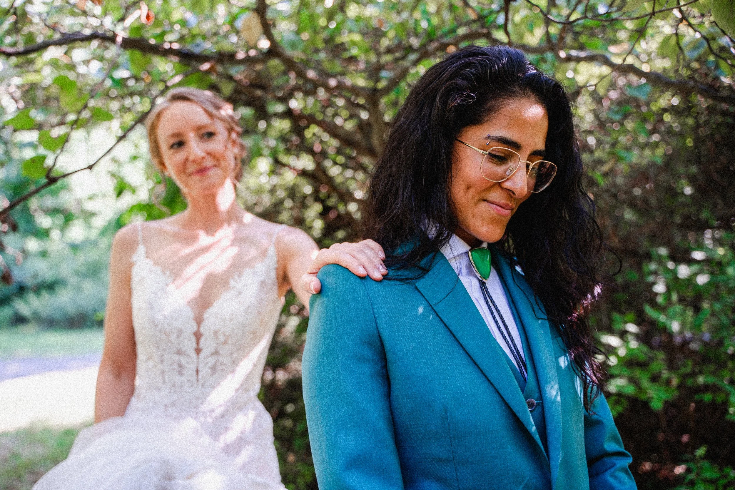 A bride with blonde hair in a white lace wedding dress places her hand on the shoulder of a woman with black curly hair and glasses, wearing a blue suit, during an outdoor wedding ceremony.