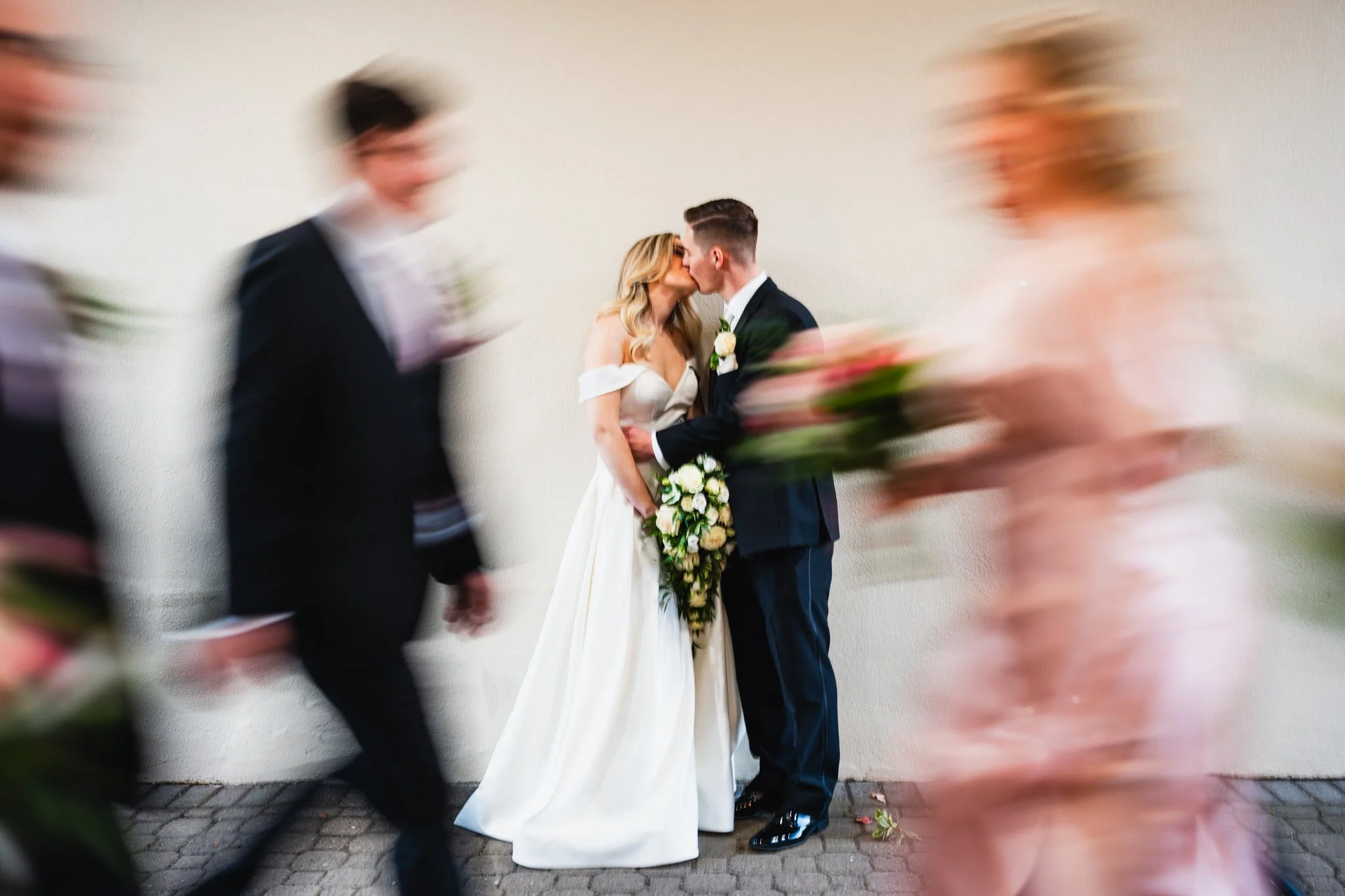 A bride and groom kiss during their wedding ceremony, surrounded by blurred wedding guests, against a plain white wall.