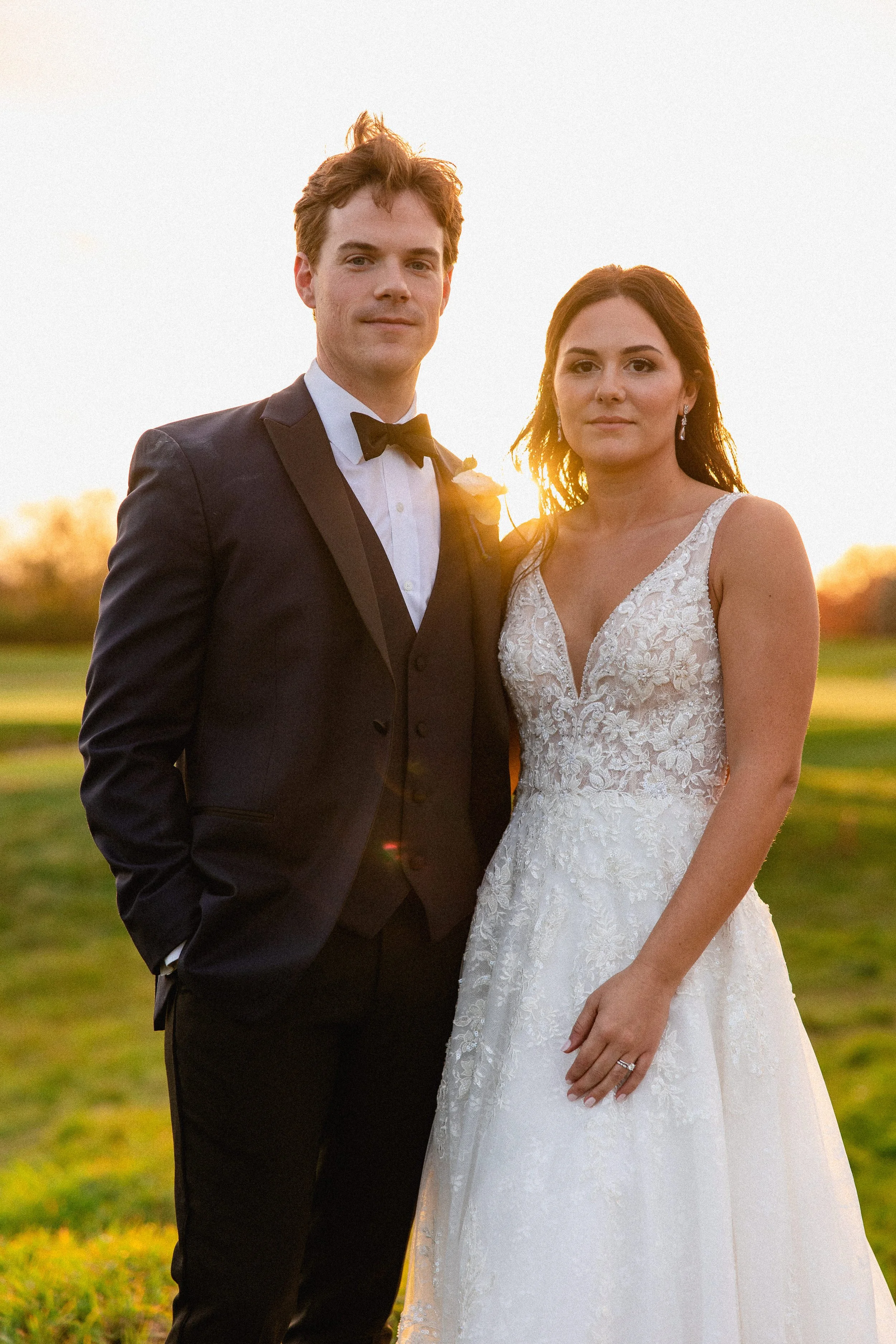 A bride and groom standing outdoors during sunset, with the groom in a black tuxedo and the bride in a white lace wedding gown.