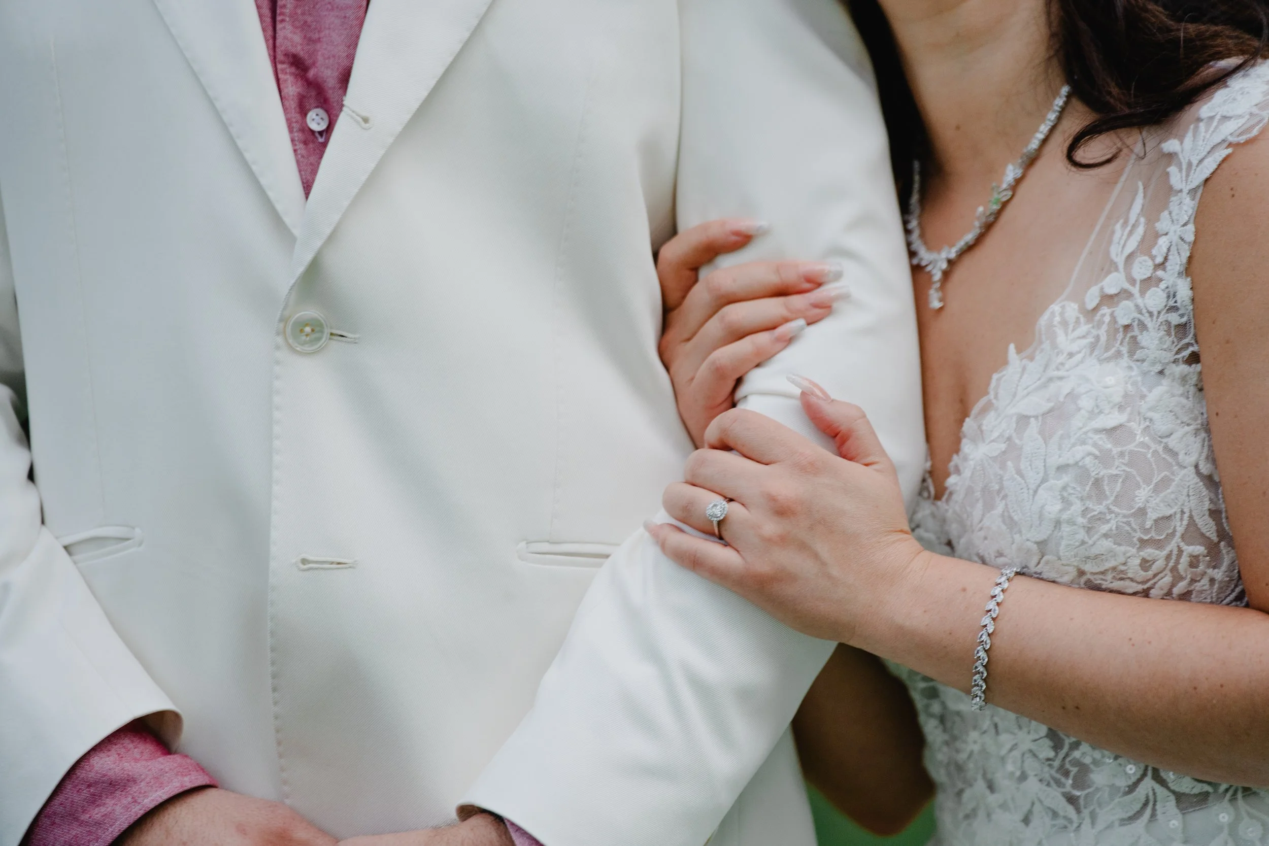 Close-up of a bride and groom holding hands, showing the bride's engagement ring, lace wedding dress, and jewelry, with the groom in a white suit and pink shirt.