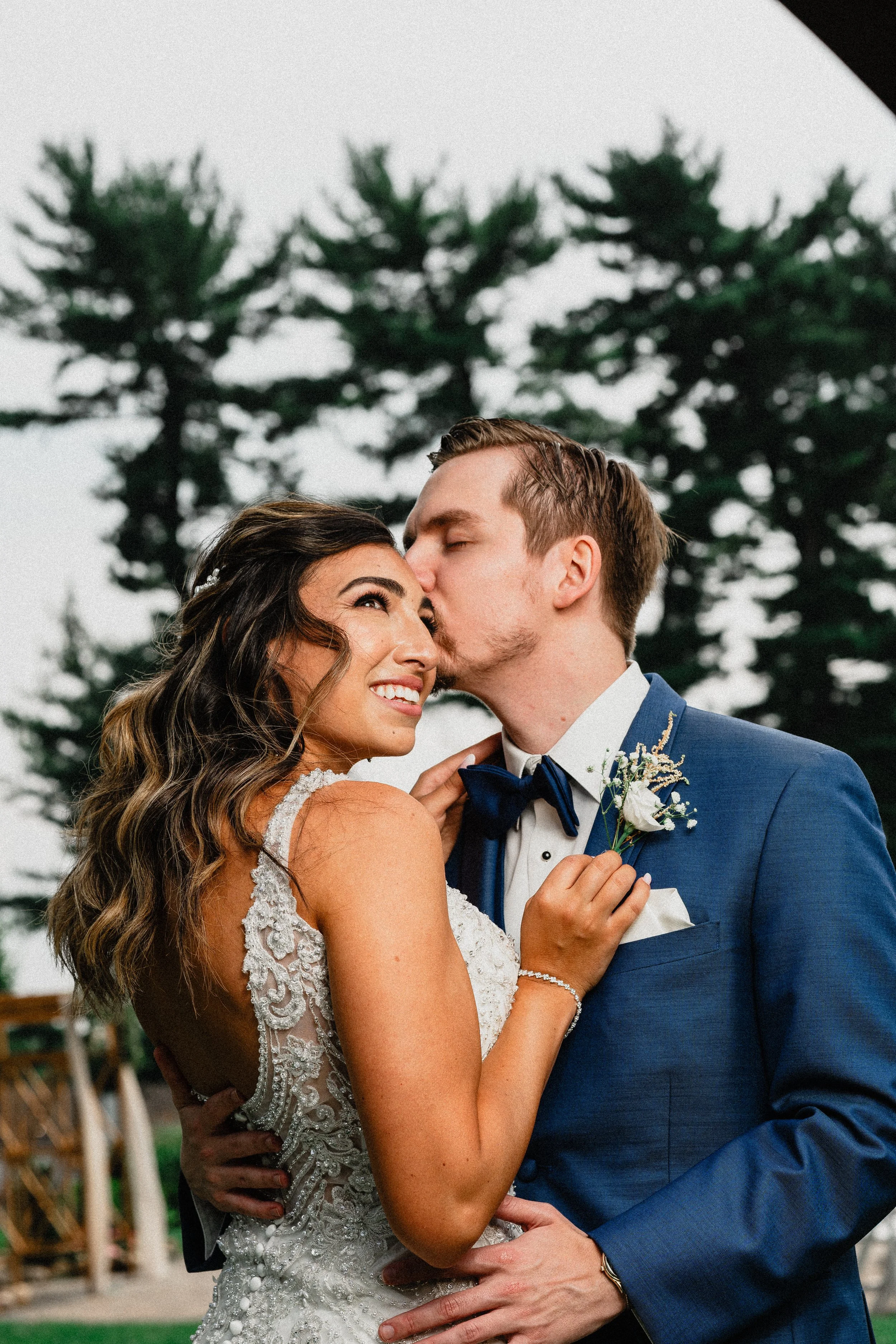 A bride and groom sharing a kiss outdoors, with the groom pressing his lips to the bride's forehead and the bride smiling, in front of a background of tall trees.