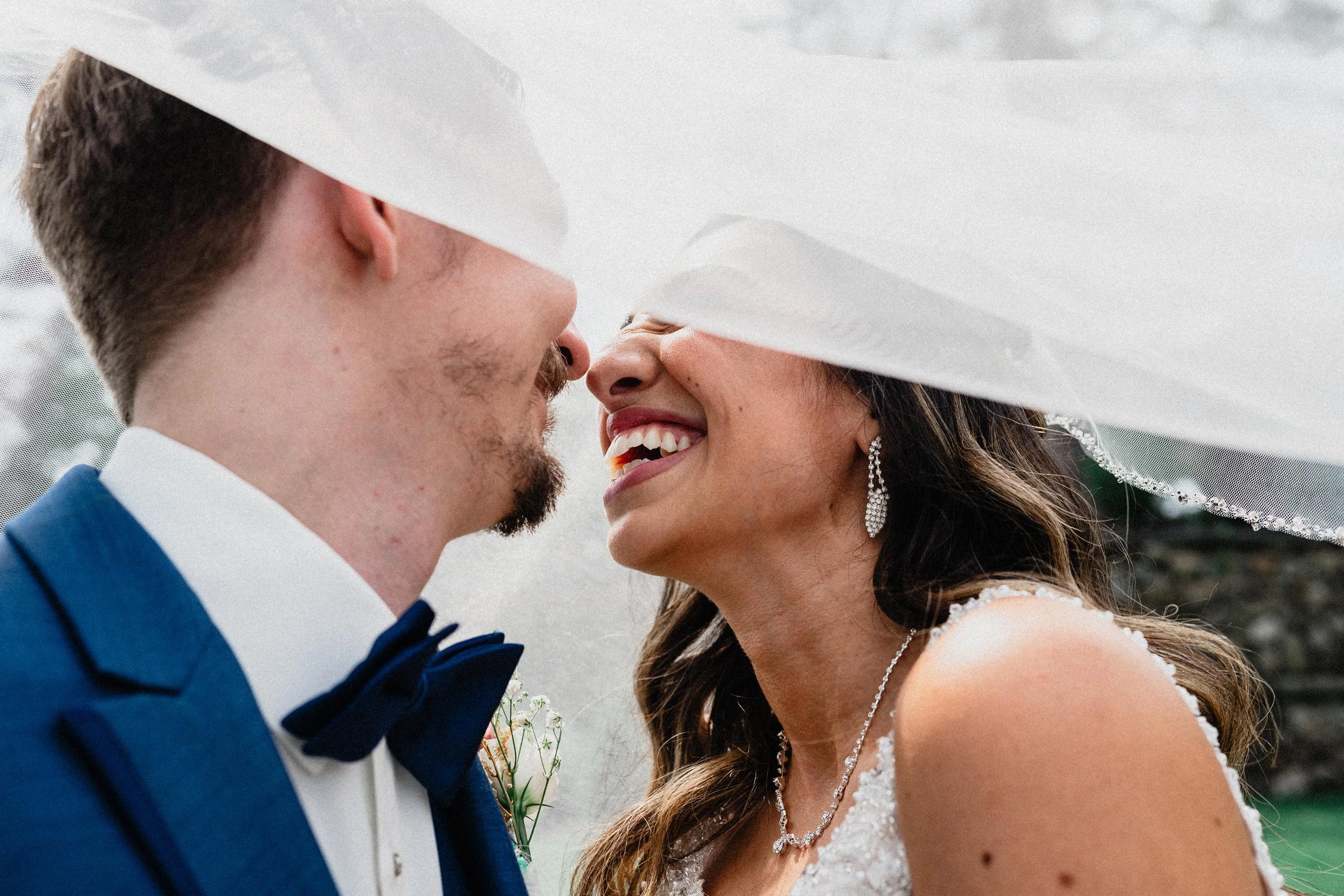 A bride and groom face close together, smiling under a wedding veil on an outdoor wedding day.