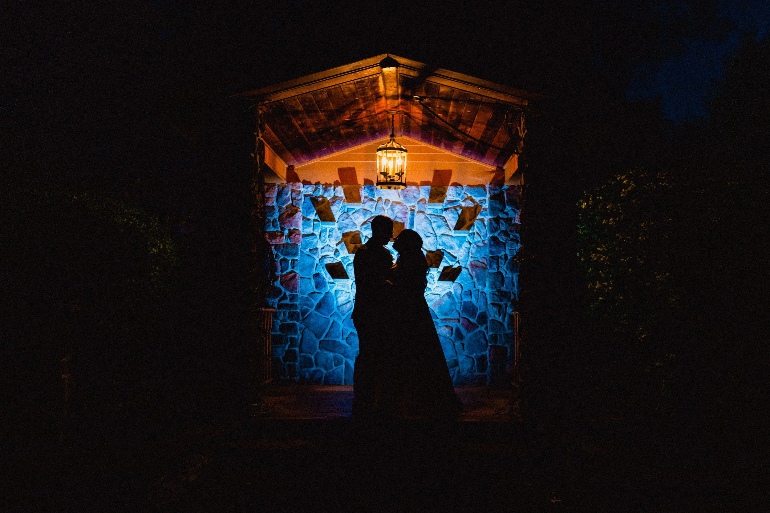 Silhouette of a couple kissing in front of a lit stone wall at night, with warm light hanging above.