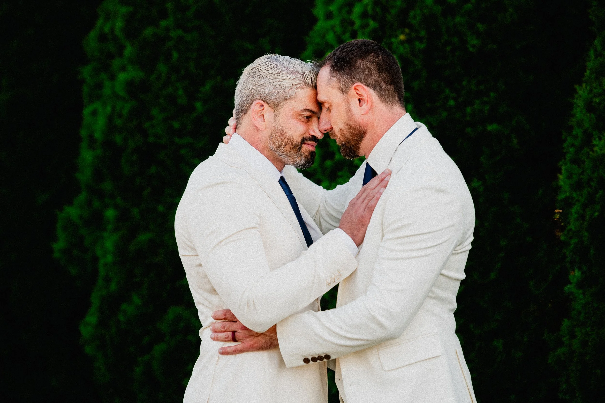 Two men in white suits with dark ties embrace and touch foreheads against a dark green, leafy background.