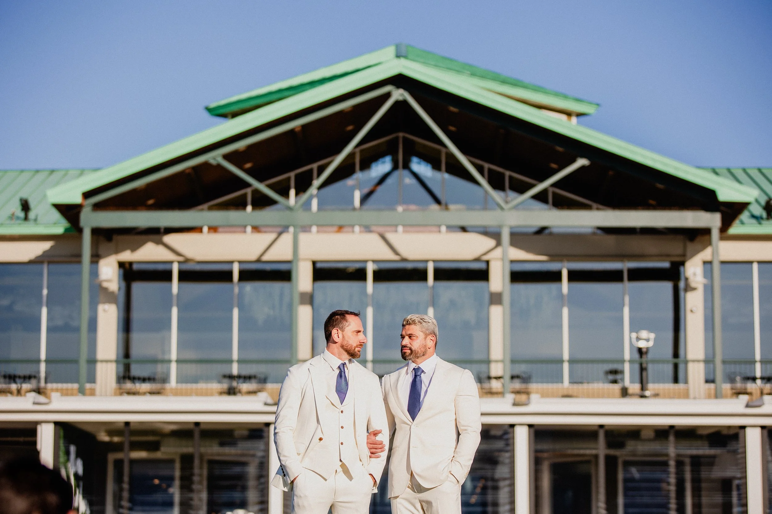 Two men in white suits and blue ties standing together outdoors in front of a large building with a green roof, engaged in conversation.