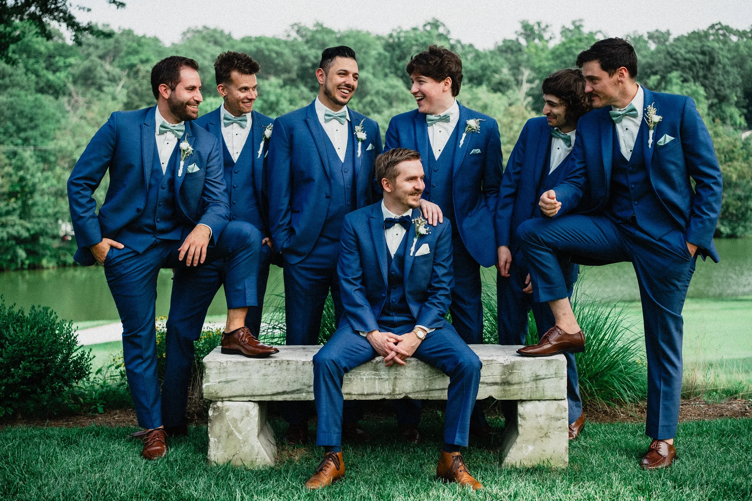 A group of eight men in blue suits and matching light blue bow ties, gathered outdoors by a pond and green trees, smiling and posing for a photo, with one seated in front and the others standing around him.