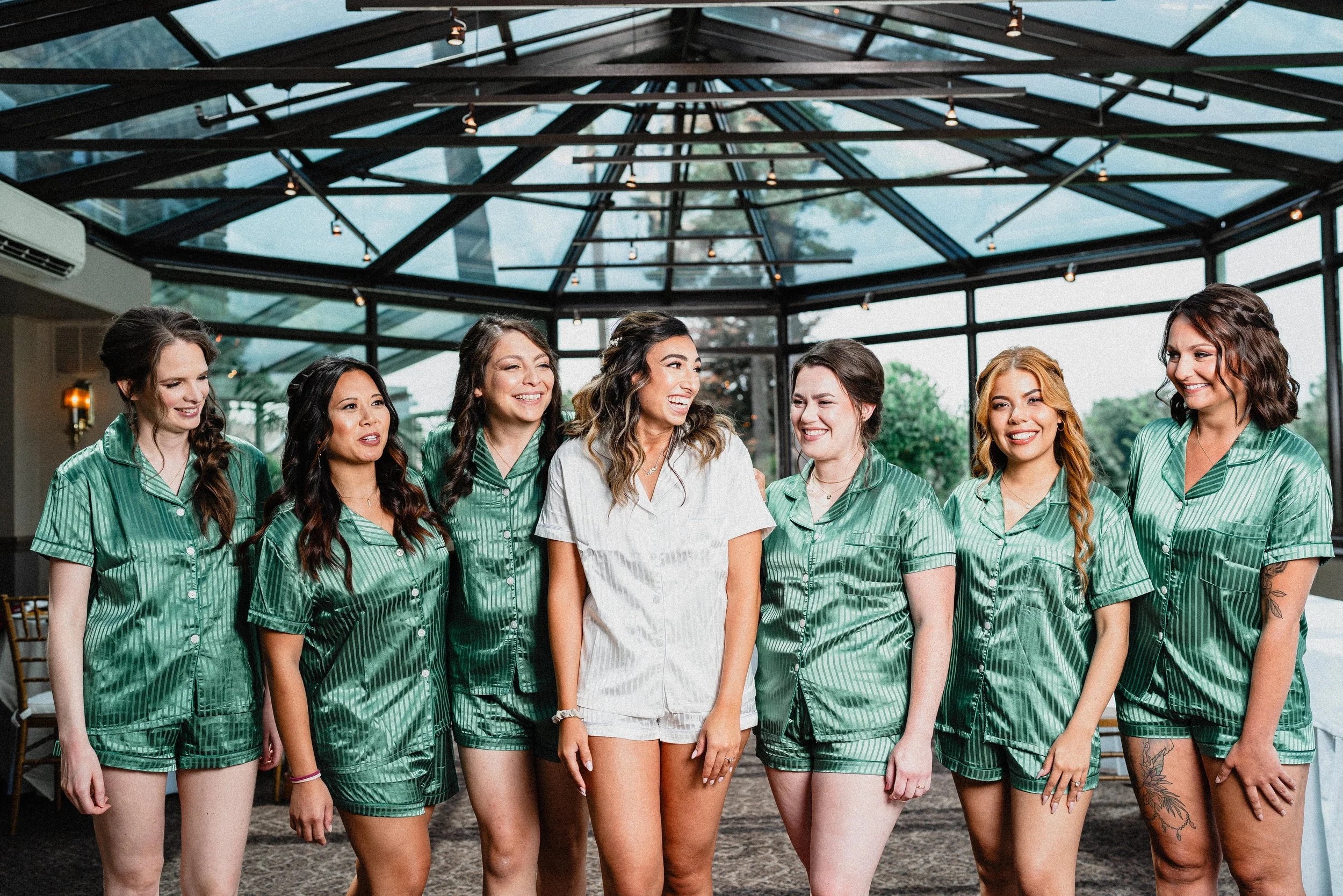 Group of seven women in matching green satin pajamas, smiling and standing together in a glass-ceilinged indoor space, celebrating a bridal shower or bachelorette party.