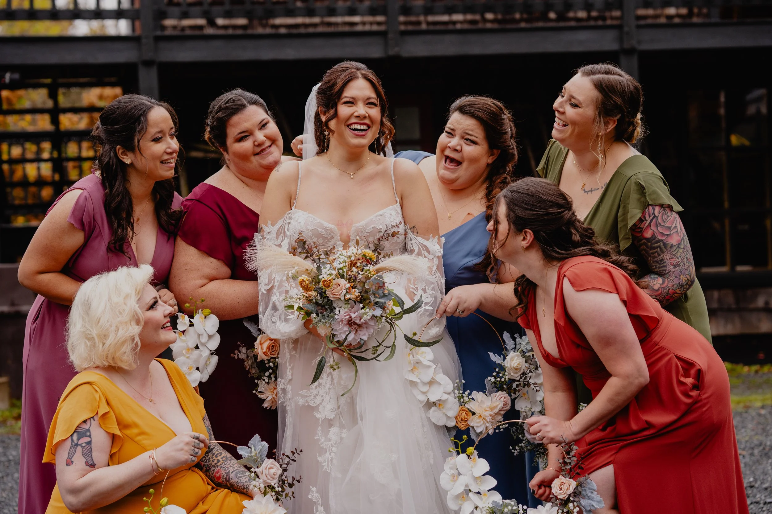 A bride in a white wedding dress and veil holding a bouquet of flowers, surrounded by seven women in colorful dresses, smiling and posing outdoors.