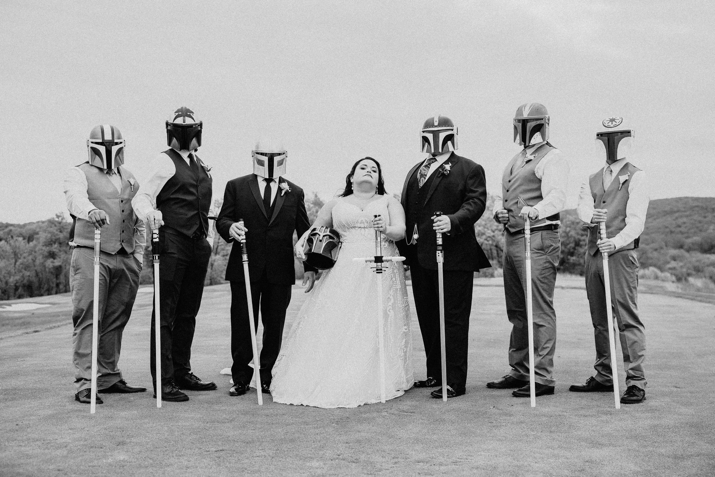 A bride in a white wedding dress standing among six men with Star Wars Mandalorian helmets, all holding canes or swords, in an outdoor setting.