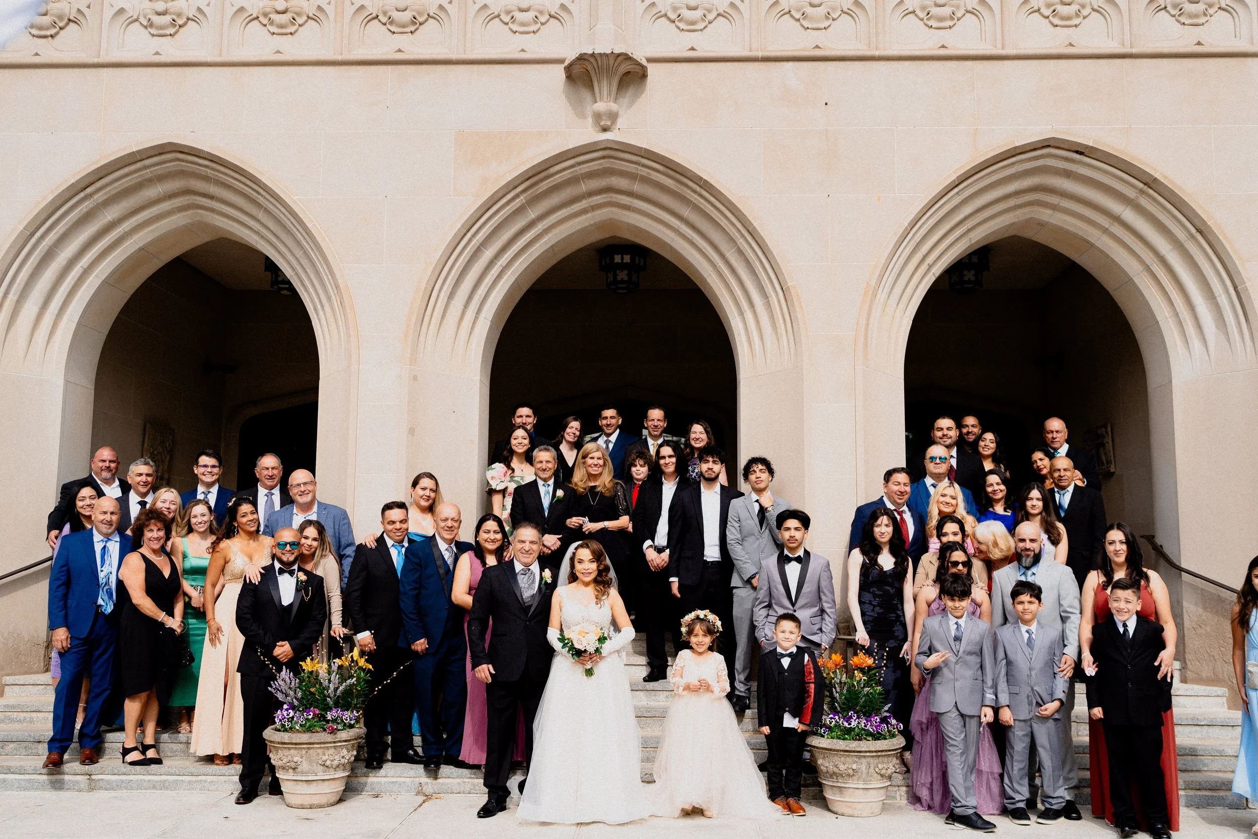 A large group of people gathered on the steps of a cream-colored building with Gothic-style arches, celebrating a wedding. The bride and groom are in the front center, smiling and holding bouquets. The group includes men, women, and children dressed 