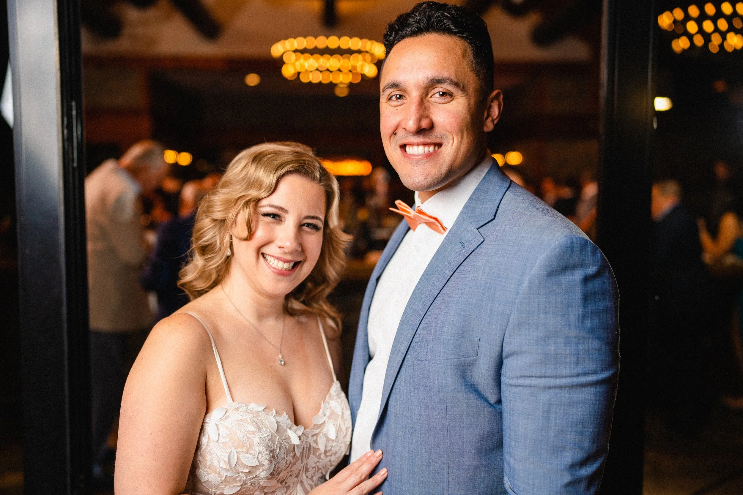 A smiling woman and man dressed in formal attire at a party or wedding reception, with warm lighting and blurred guests in the background.
