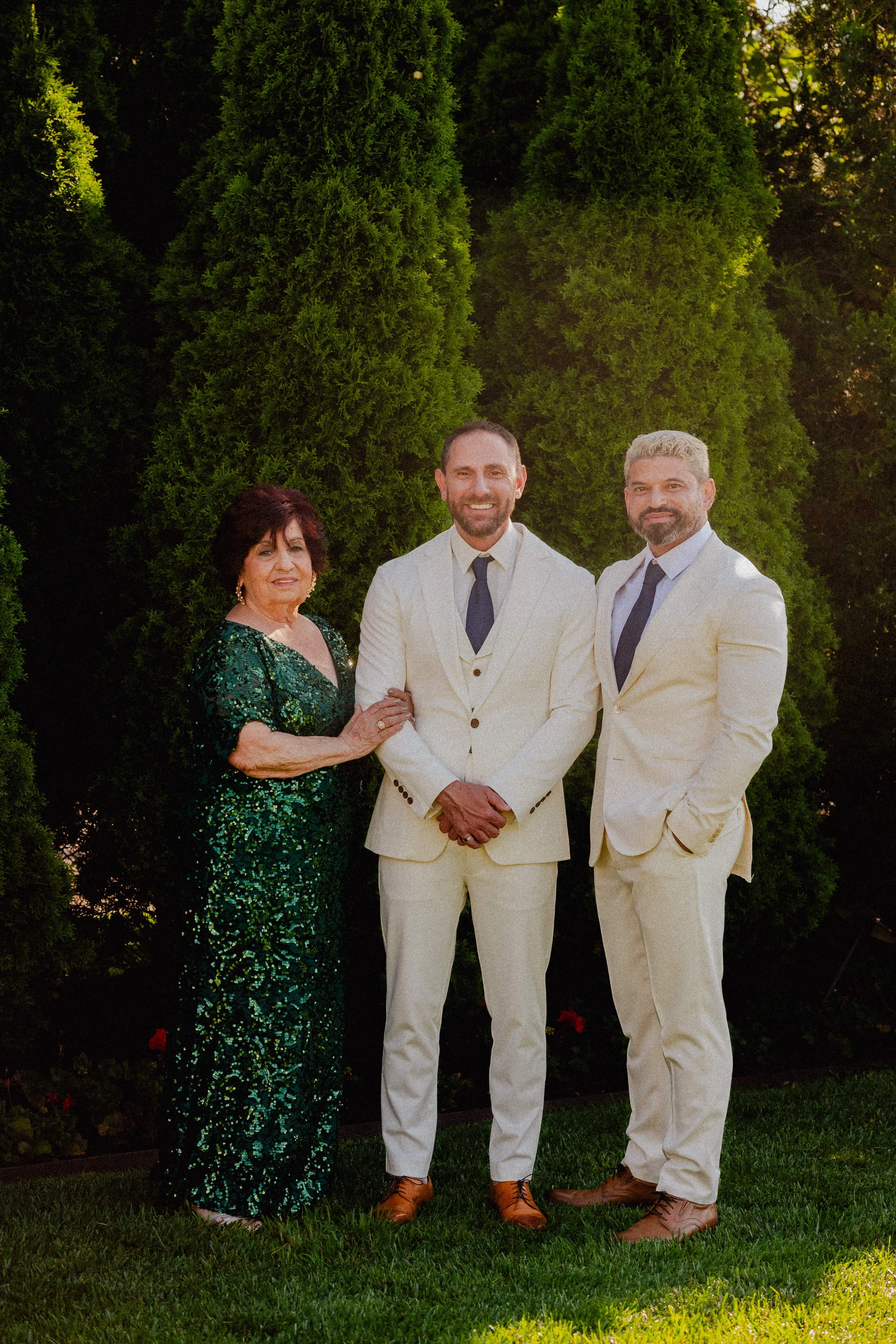 Three people dressed in formal attire standing outdoors in front of tall green trees. One woman wearing a dark green sequined gown and two men in white suits with dark ties.