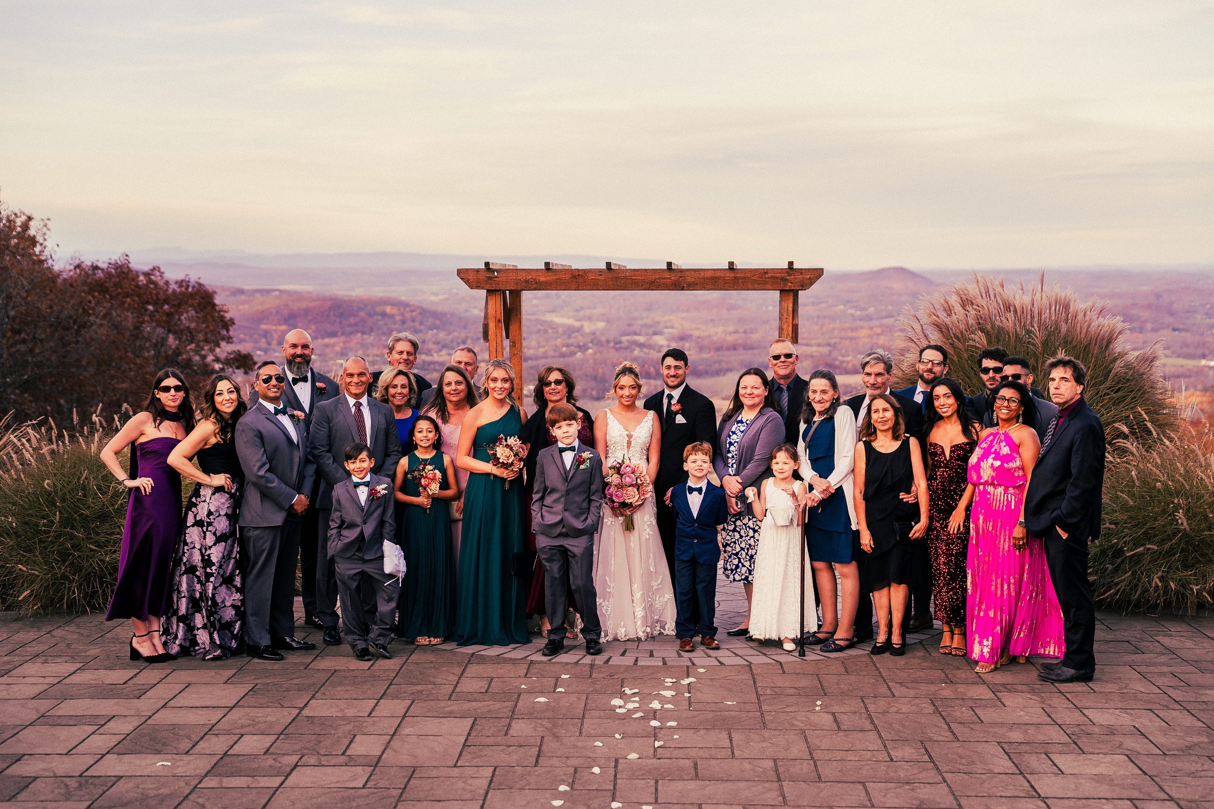 Group of wedding guests and family members gathered outdoors, dressed in formal attire, posing for a group photo with a scenic landscape in the background.