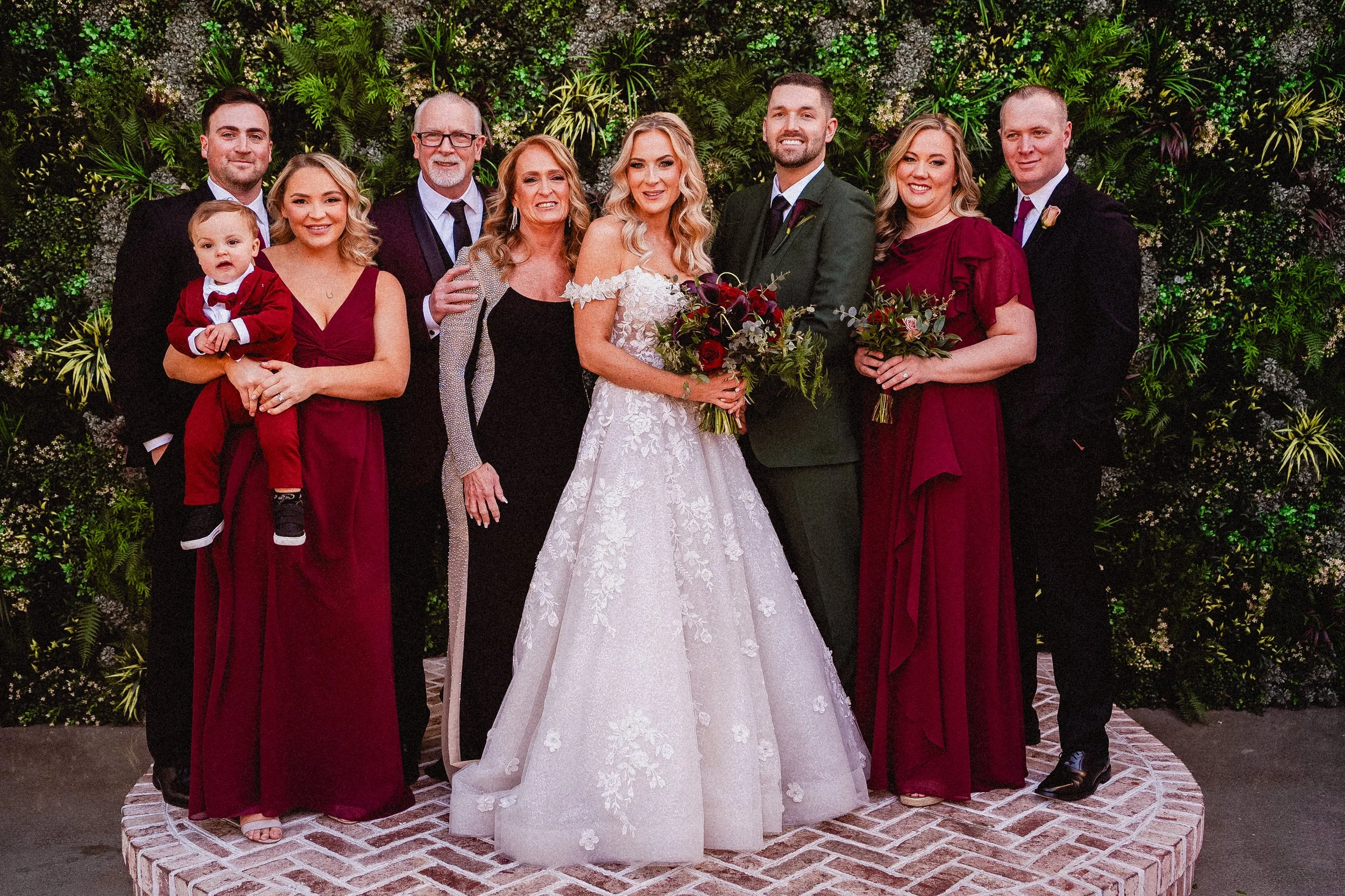 Group of people at a wedding, including bride in white gown, groom in green suit, and family members, standing on a circular brick platform in front of a lush green floral backdrop.