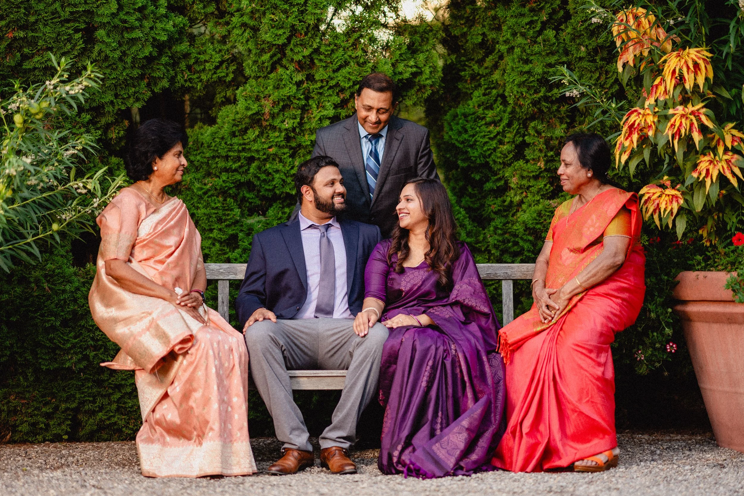 A family of six Indian people sitting on a garden bench, smiling and looking at each other, with lush green bushes and potted plants in the background.